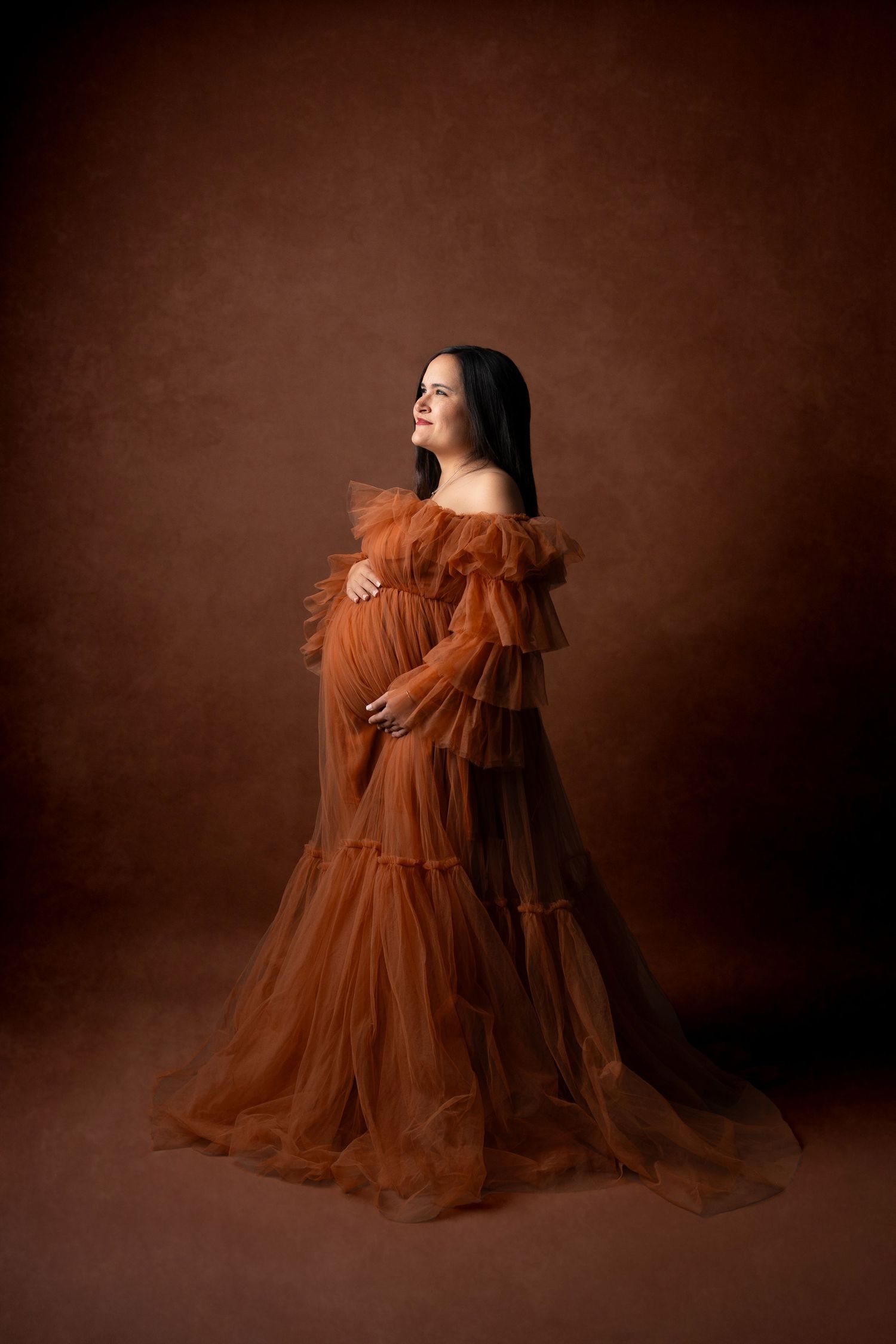 Ethereal maternity portrait in flowing rust-colored tulle gown against dark background.