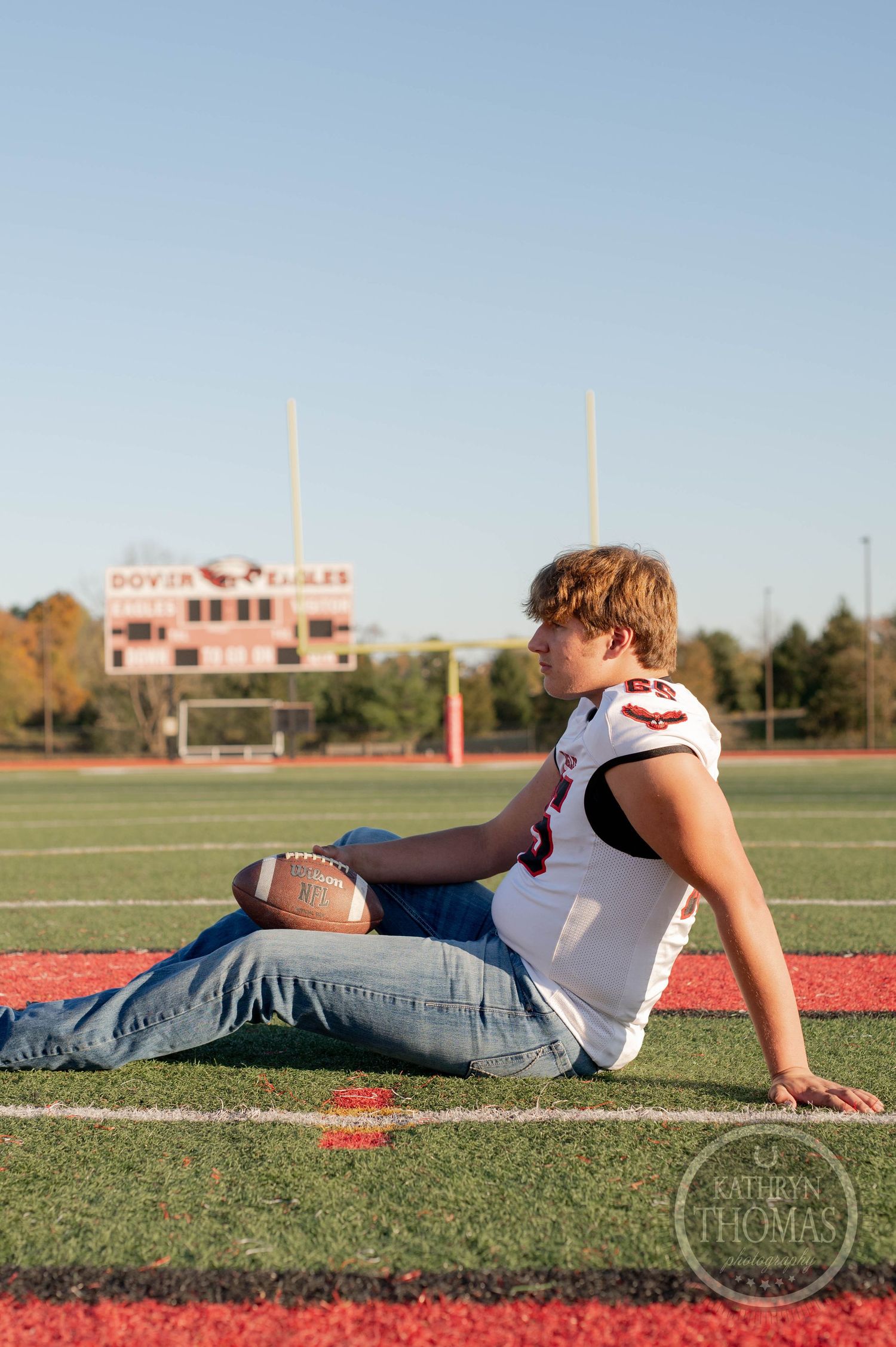 Athlete in white jersey sits casually on field with football and scoreboard visible in background.