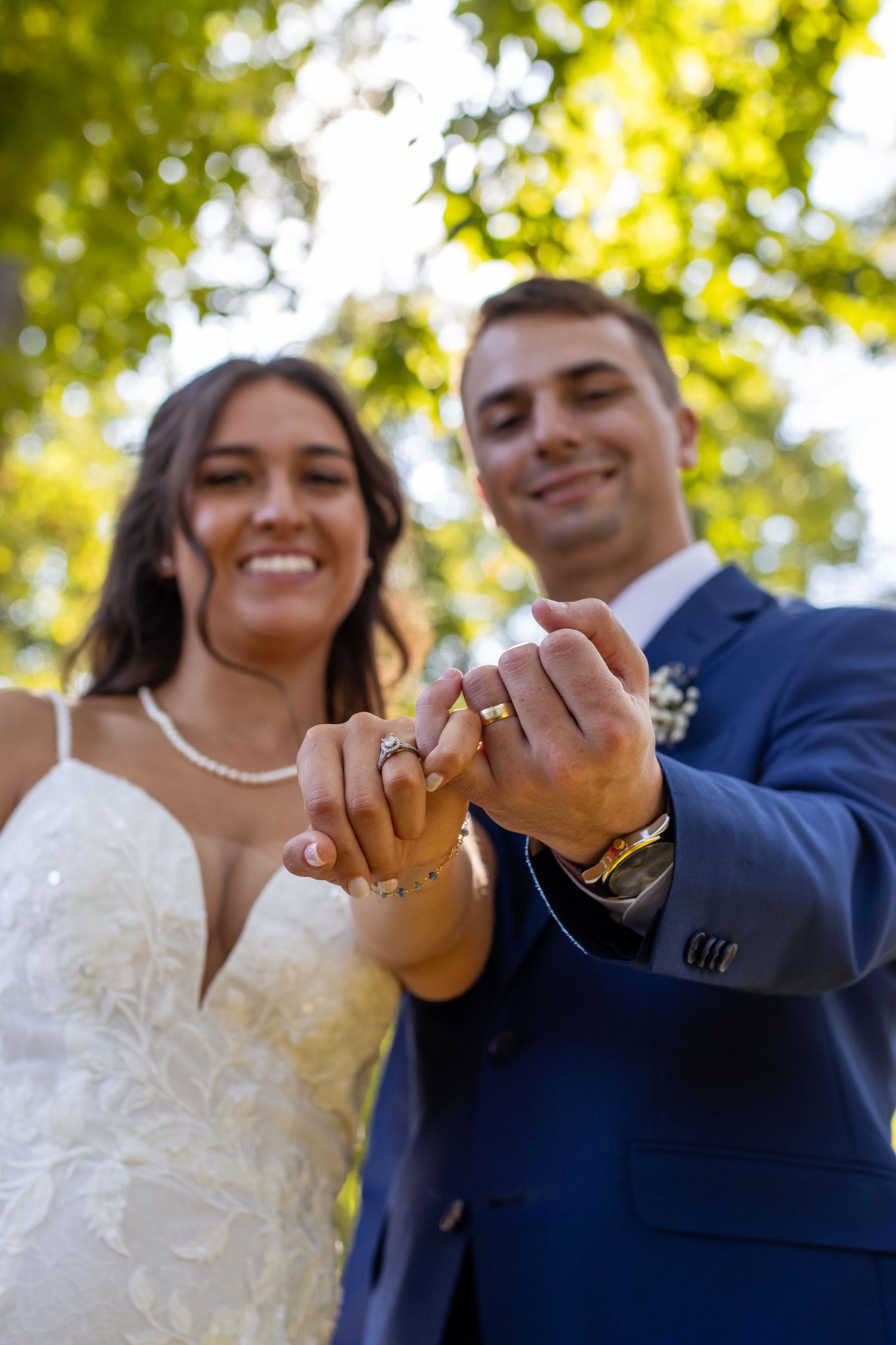 Happy newlyweds show off their wedding rings outside on a sunny day beneath green trees.