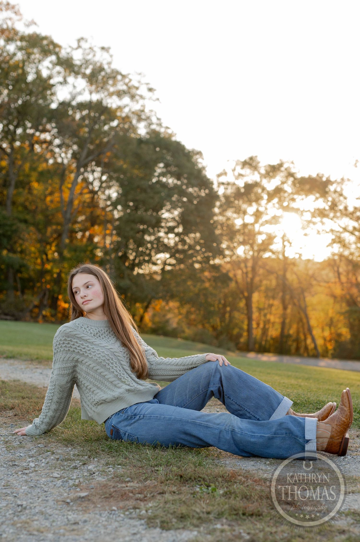 Person reclining on gravel path wearing cable knit sweater and jeans during fall sunset.