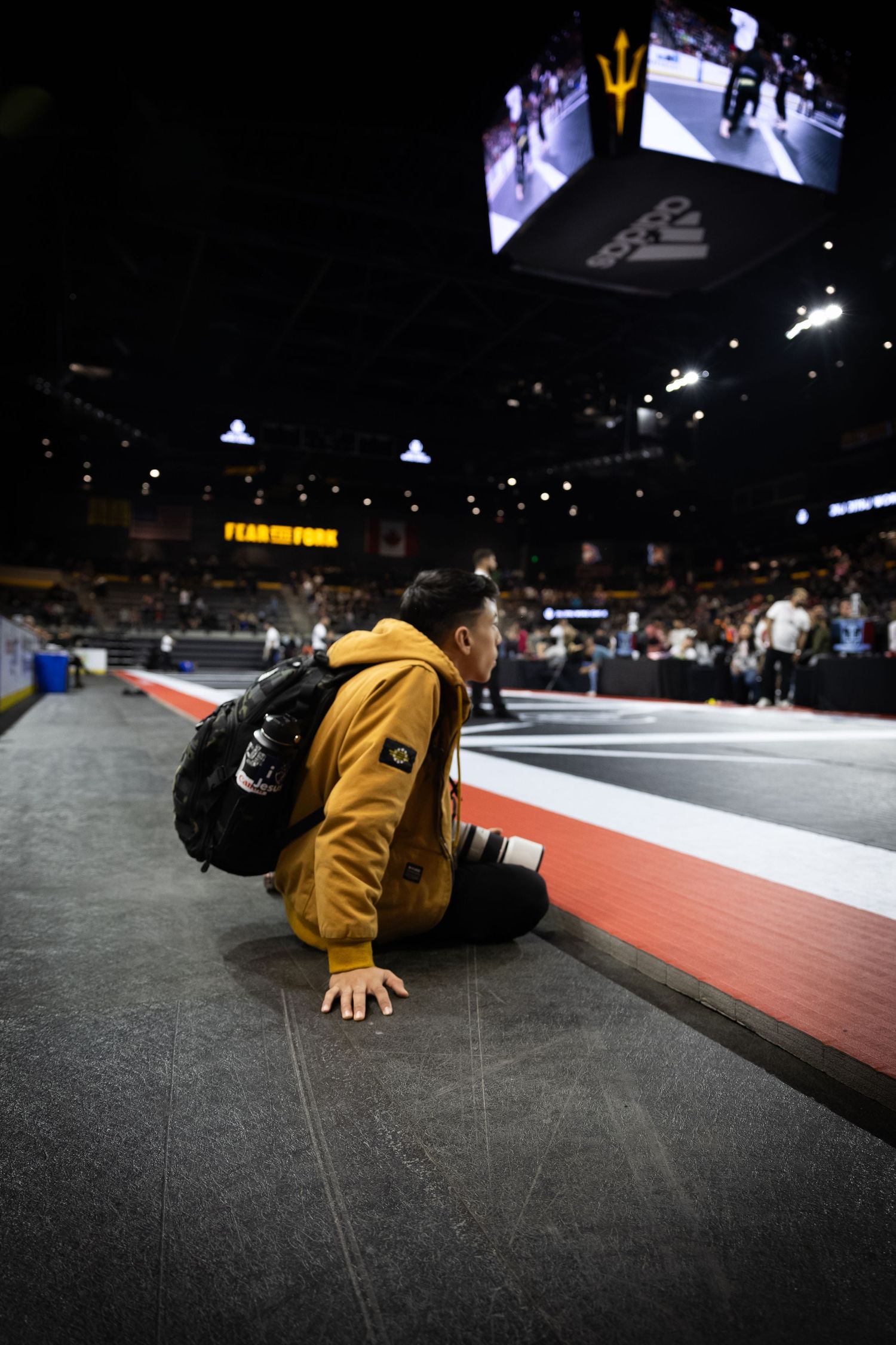 Brazilian jiu jitsu photographer in yellow jacket sits at edge of competition mat with his camera ready to shoot the action shots