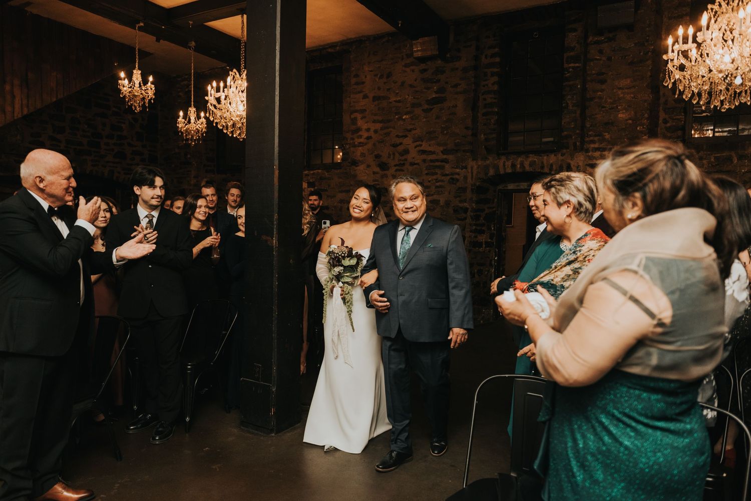 Wedding ceremony in a rustic venue with brick walls, chandeliers, and guests gathered to celebrate the couple's entrance.