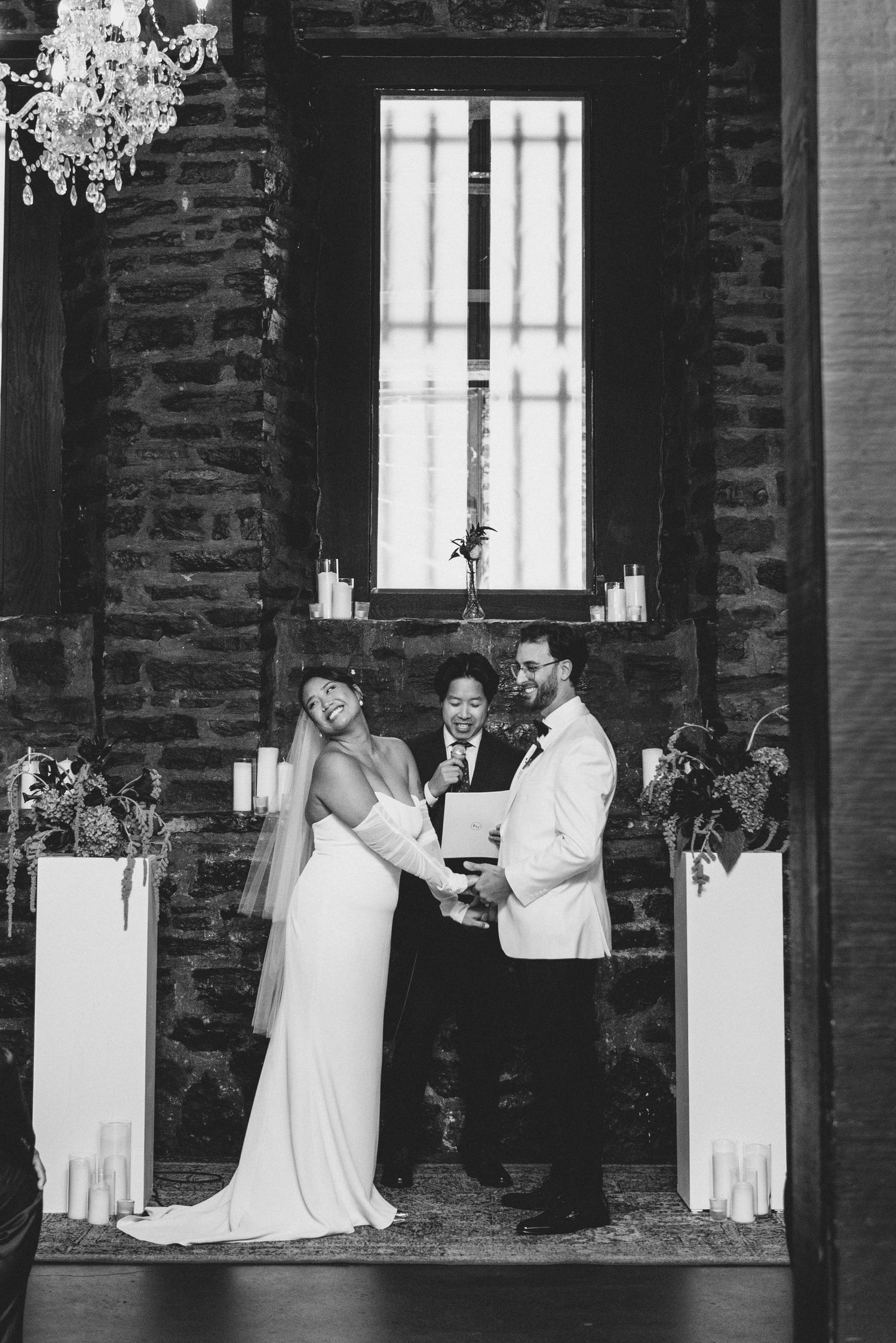 Black and white wedding ceremony inside a rustic brick venue with chandelier and candlelit decor.