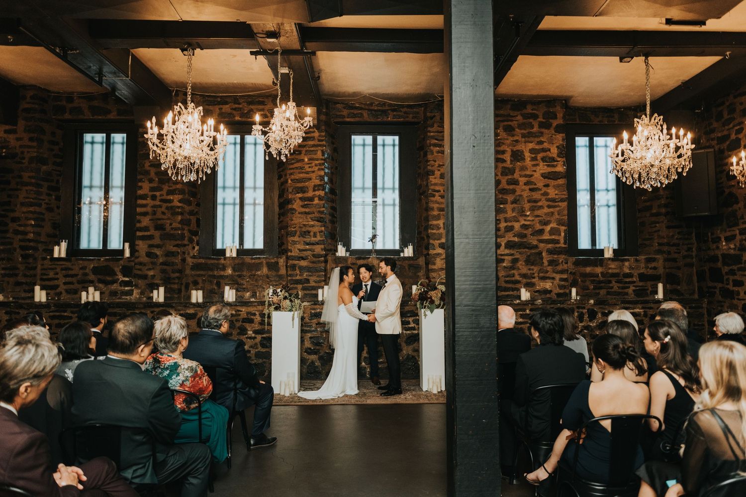 A wedding ceremony in a rustic brick venue with elegant crystal chandeliers and guests seated in rows.