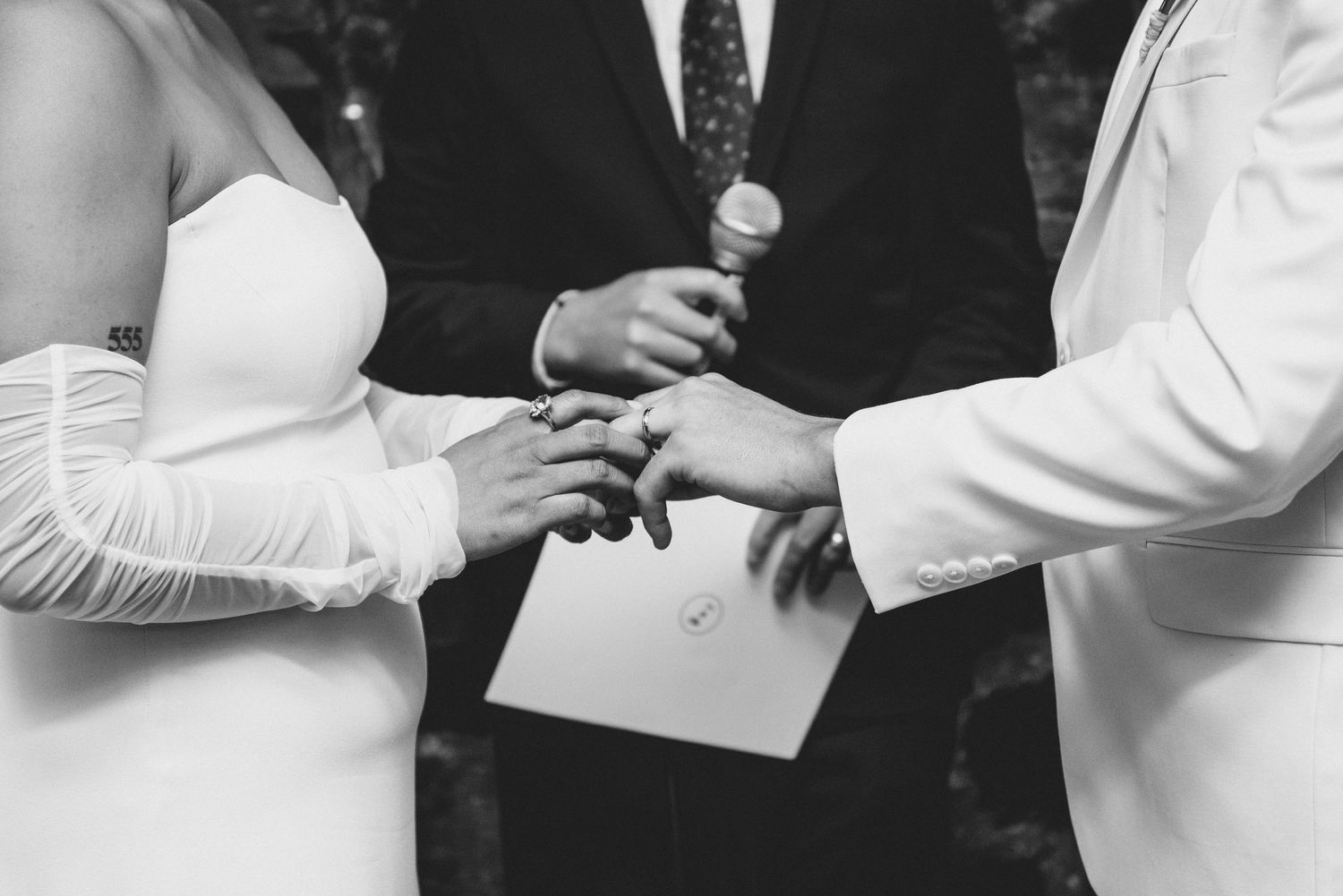 Close-up black and white image of exchanging wedding rings during a ceremony with white attire visible.