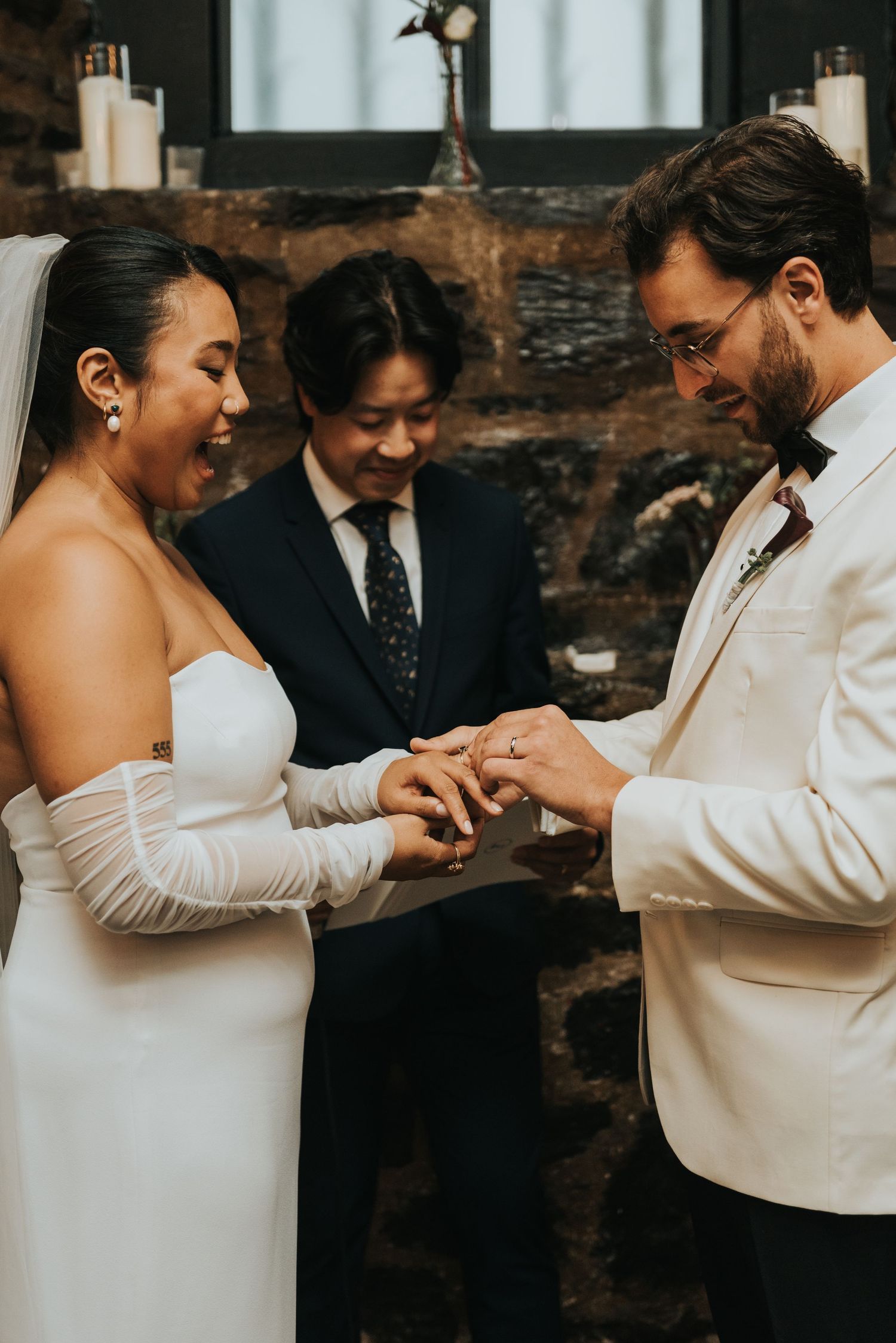 An intimate wedding ceremony showing a couple exchanging rings with an officiant in front of rustic stone wall decor.