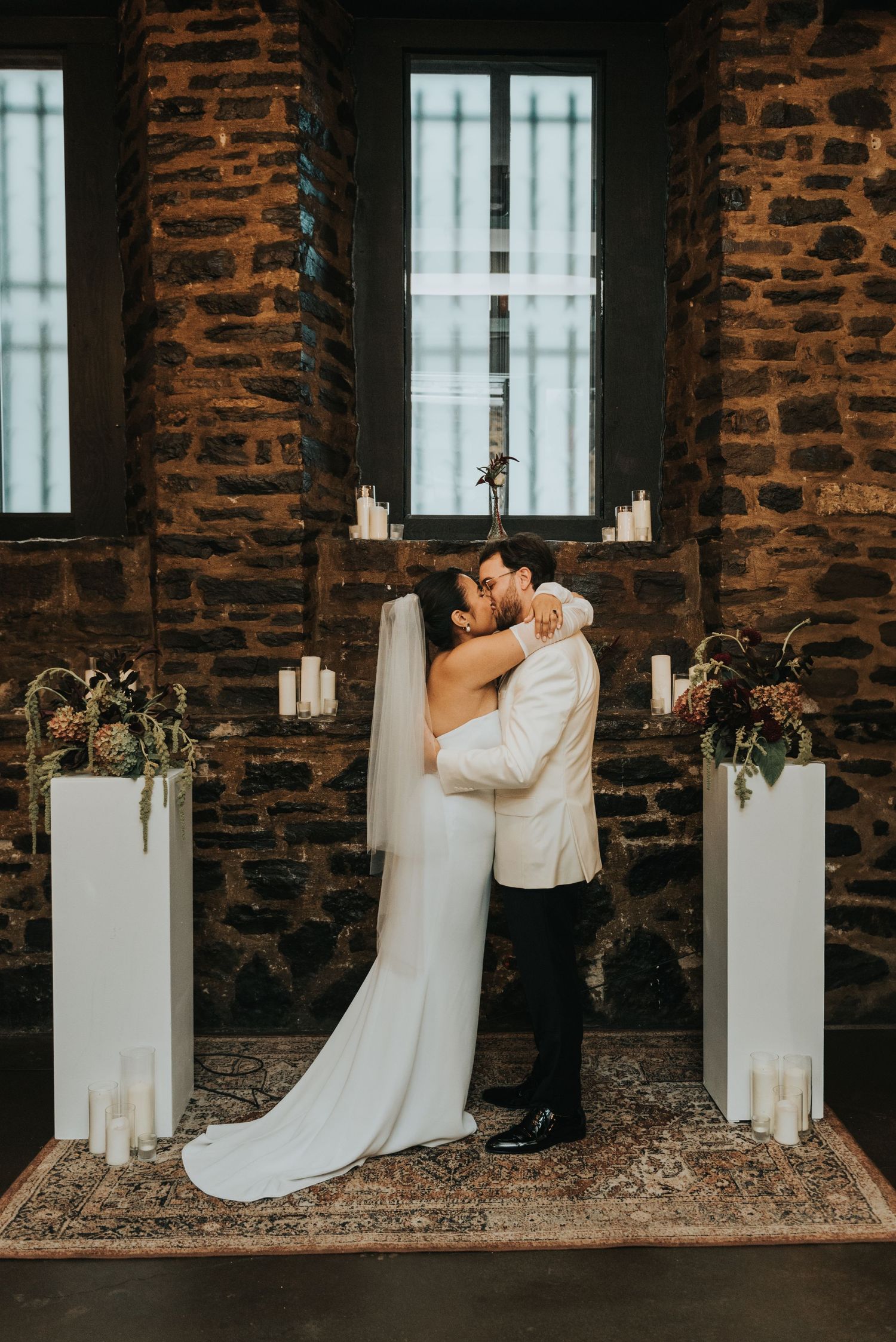 A romantic wedding ceremony setup against exposed brick walls with white pedestals, candles and floral arrangements.