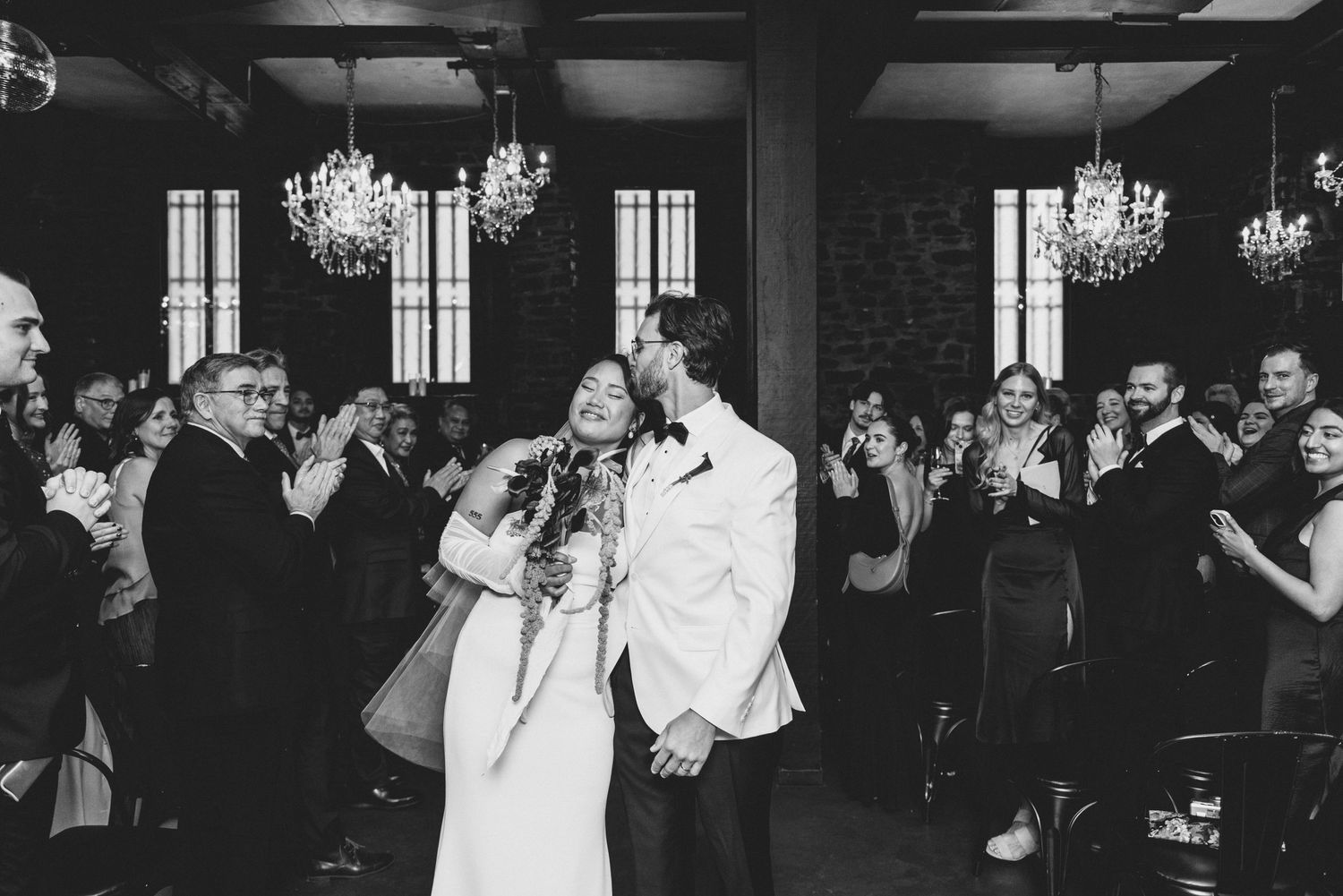 A black and white wedding celebration photo showing a couple's first dance under elegant chandeliers in a dark venue.