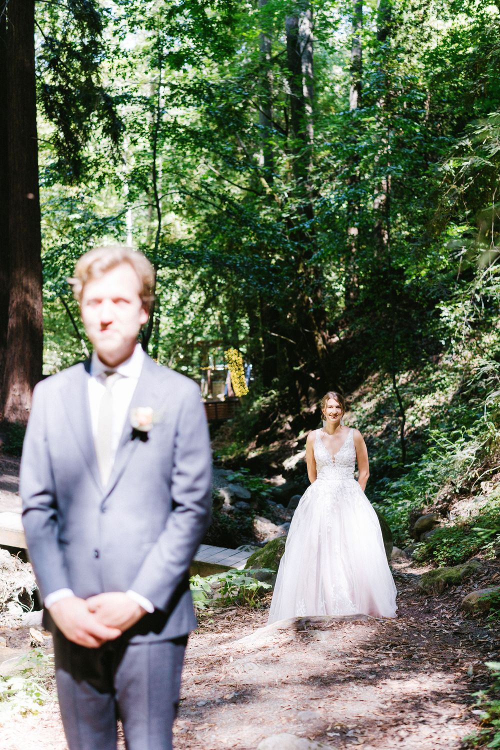 A romantic first look moment in a forest setting where a bride in white waits behind her groom.