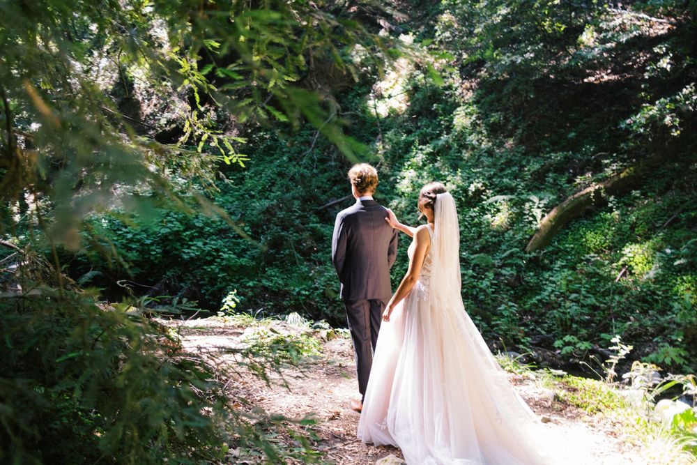 A couple in formal wedding attire walks hand in hand down a sunlit forest path surrounded by lush green foliage.