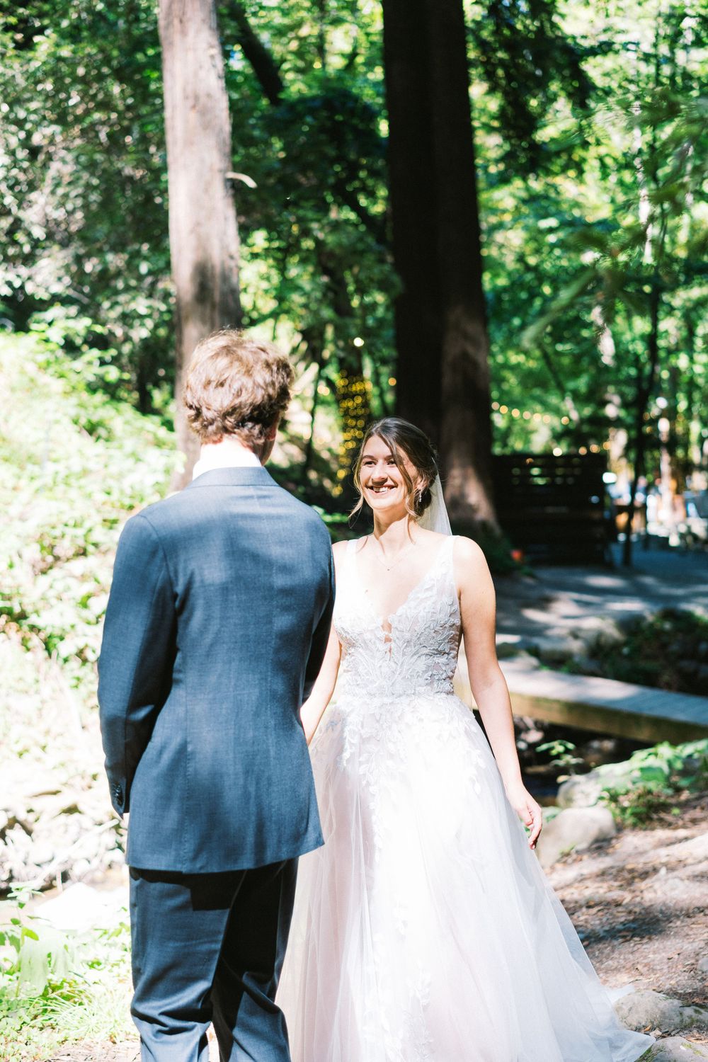 Bride in white lace gown shares a moment with groom in navy suit on a wooded path.