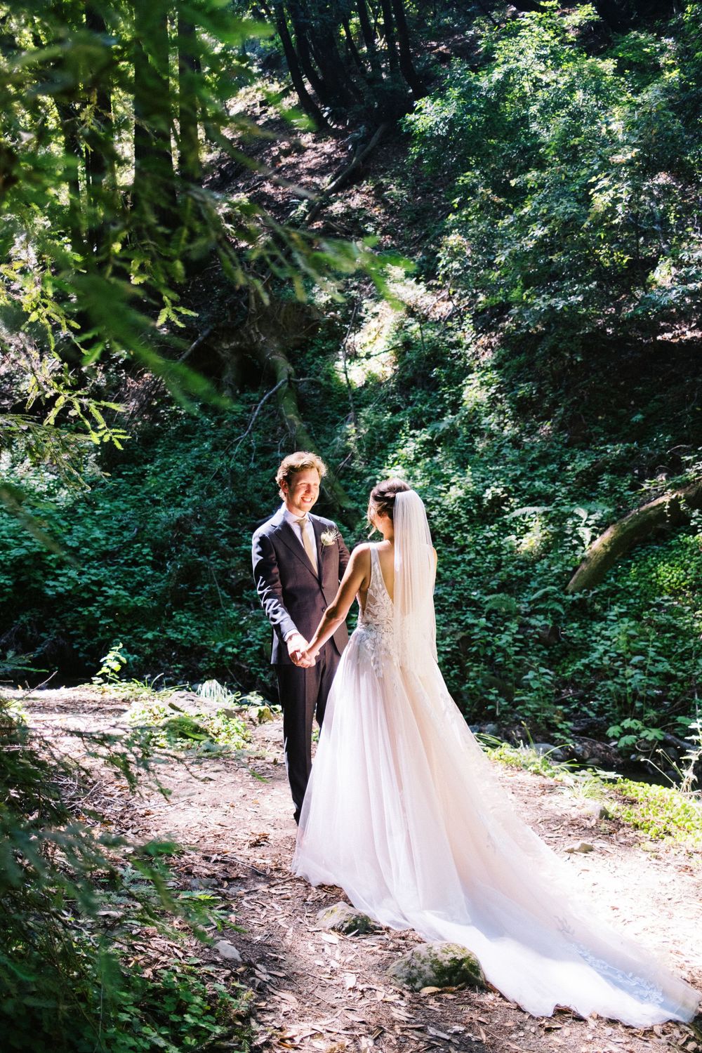 Newlyweds standing on forest path with bride wearing flowing white dress with long veil.