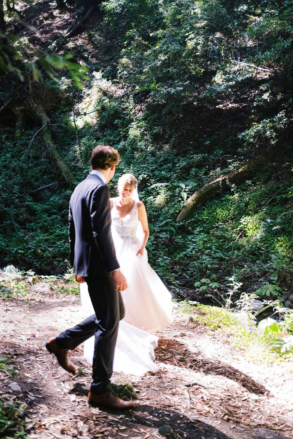 Couple in wedding attire walking together on a forest trail surrounded by lush greenery.