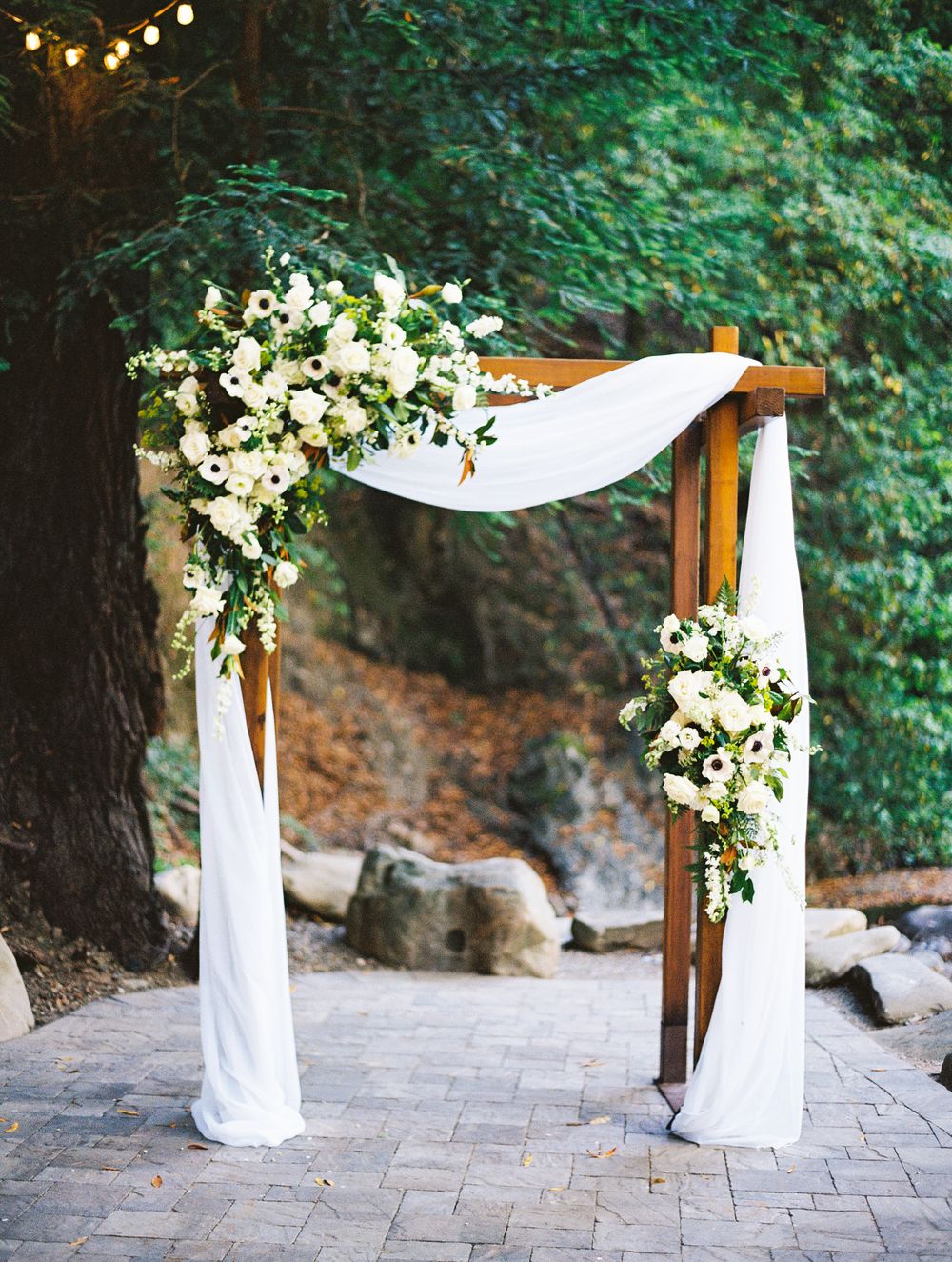 Wooden wedding arch decorated with white fabric draping and floral arrangements on a stone patio under string lights.
