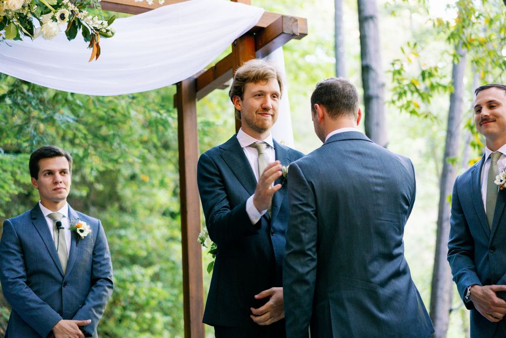 Wedding ceremony taking place outdoors under a wooden arch with groomsmen in navy suits watching the proceedings.