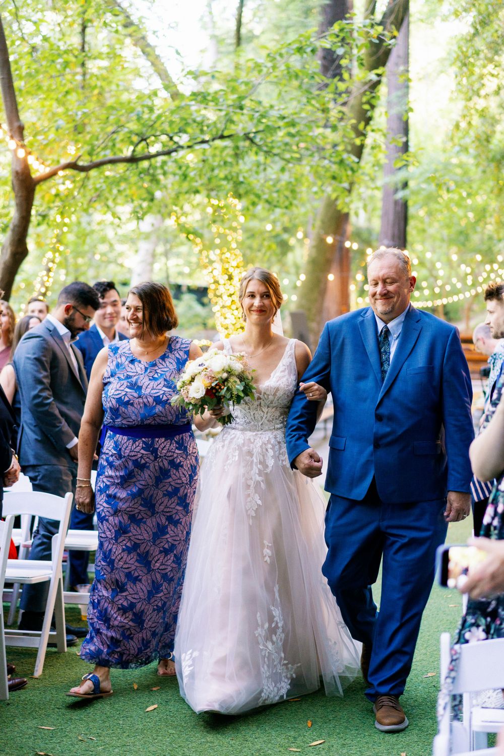 An outdoor wedding ceremony with guests seated on white chairs beneath trees decorated with string lights.