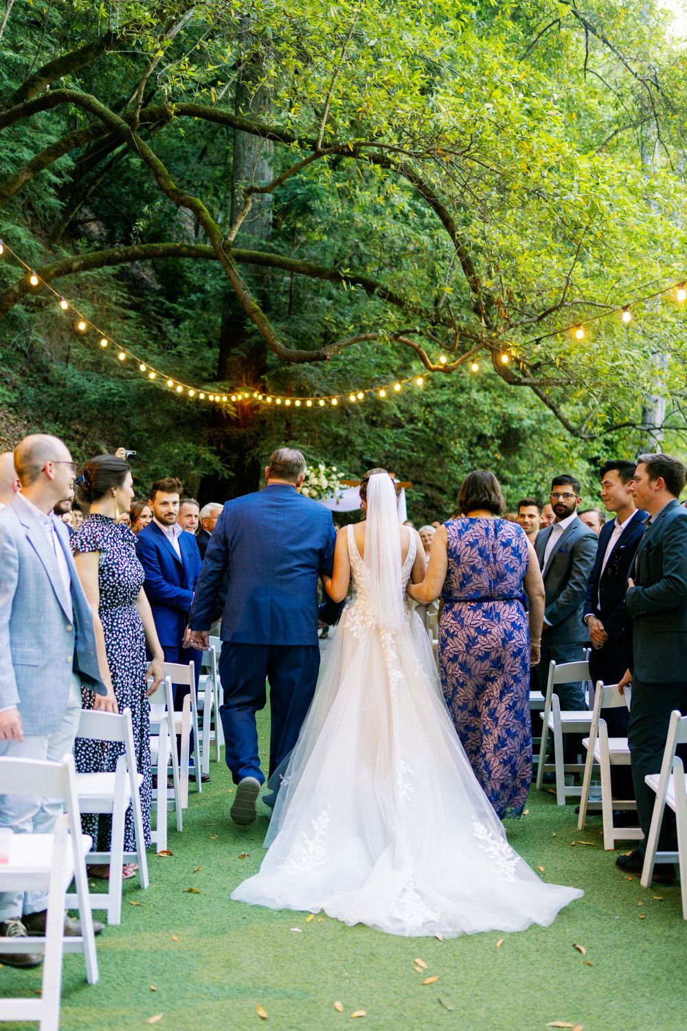 Wedding ceremony gathering on green lawn under trees with string lights overhead.