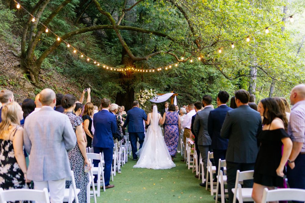 An outdoor wedding ceremony under string lights and trees with guests seated in white chairs on a grass lawn.