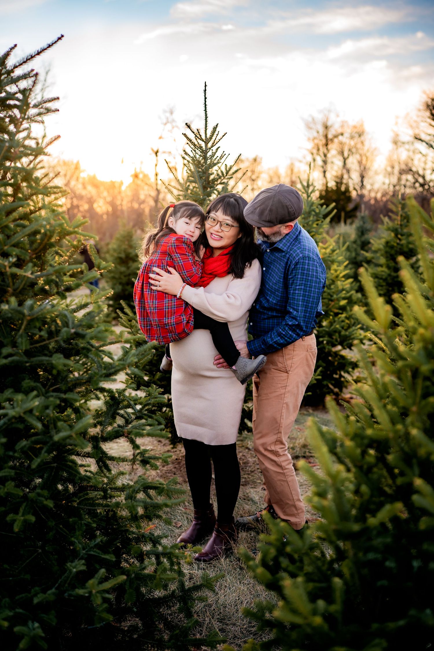 A family shares a tender moment among evergreen trees at sunset in a Christmas tree farm.