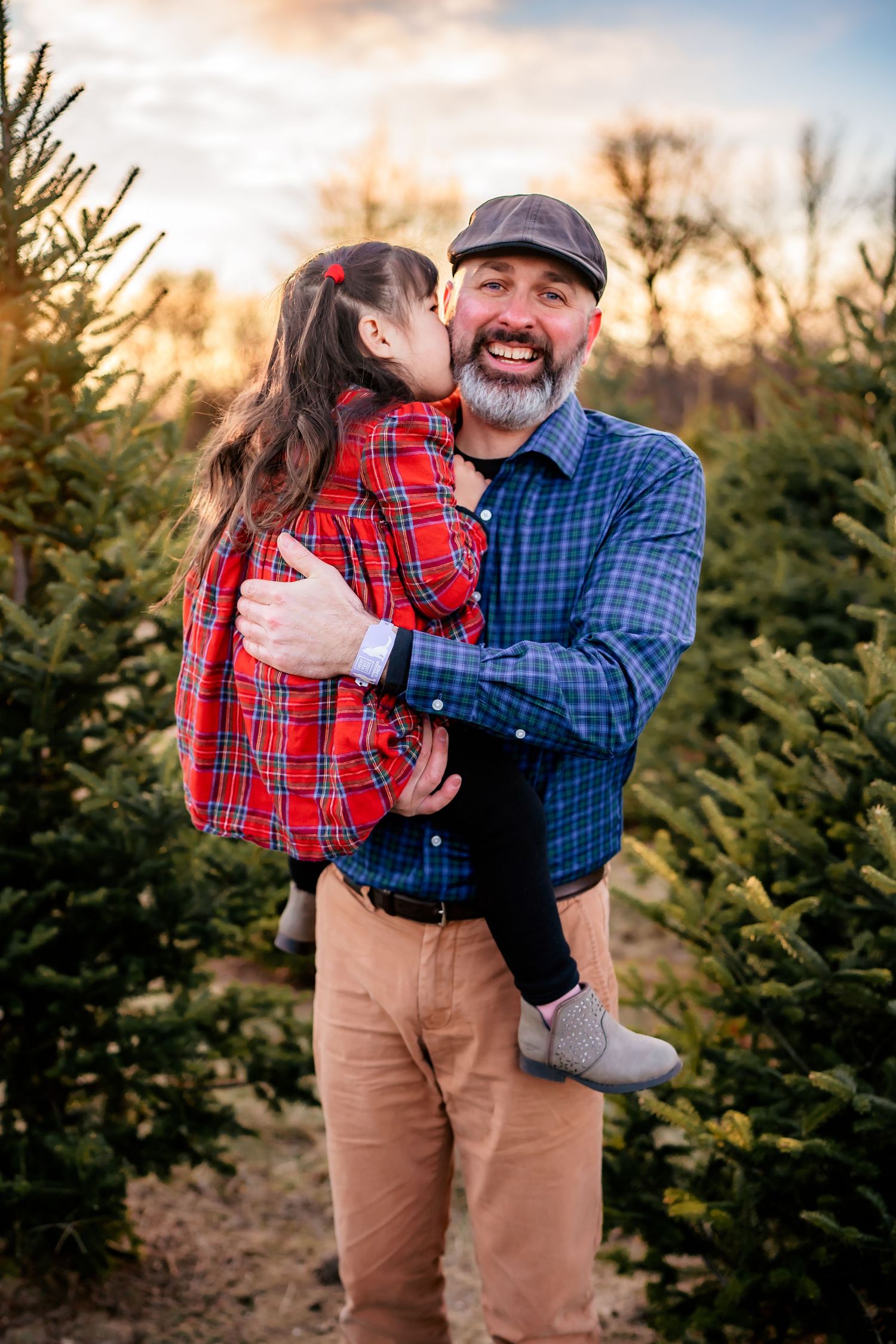 Sweet moment at a Christmas tree farm with matching red and blue plaid flannel shirts among evergreen trees at sunset.