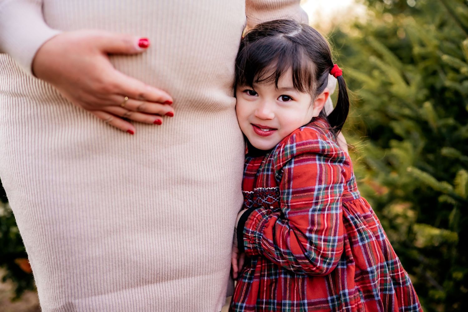 A child in a red plaid dress hugs close to someone wearing a beige sweater outdoors near greenery.