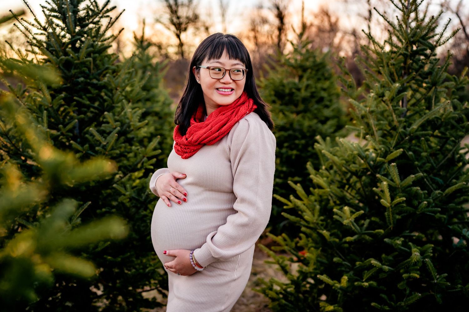 An expectant figure in a cream sweater and bright red scarf poses among evergreen trees at sunset.