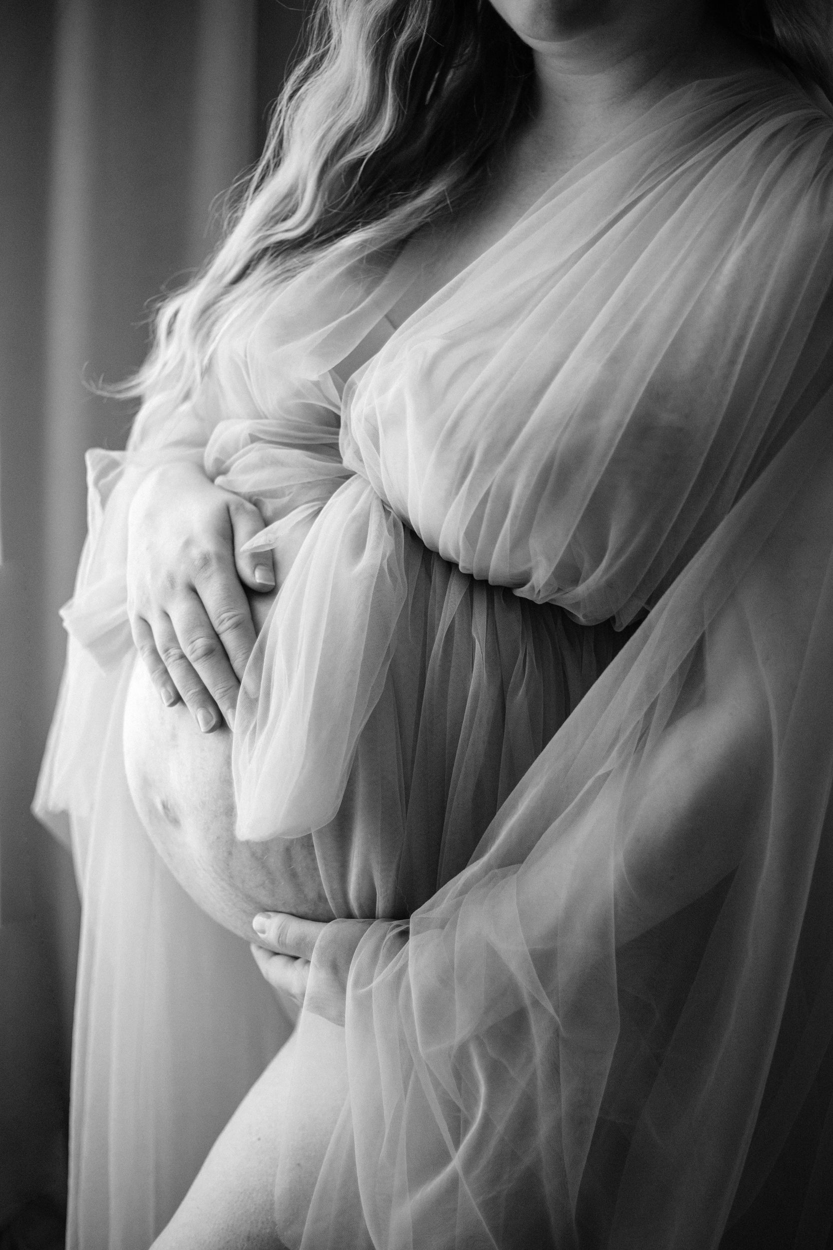 A delicate black and white maternity photo showing a woman in flowing tulle gown with hands cradling her pregnant belly.