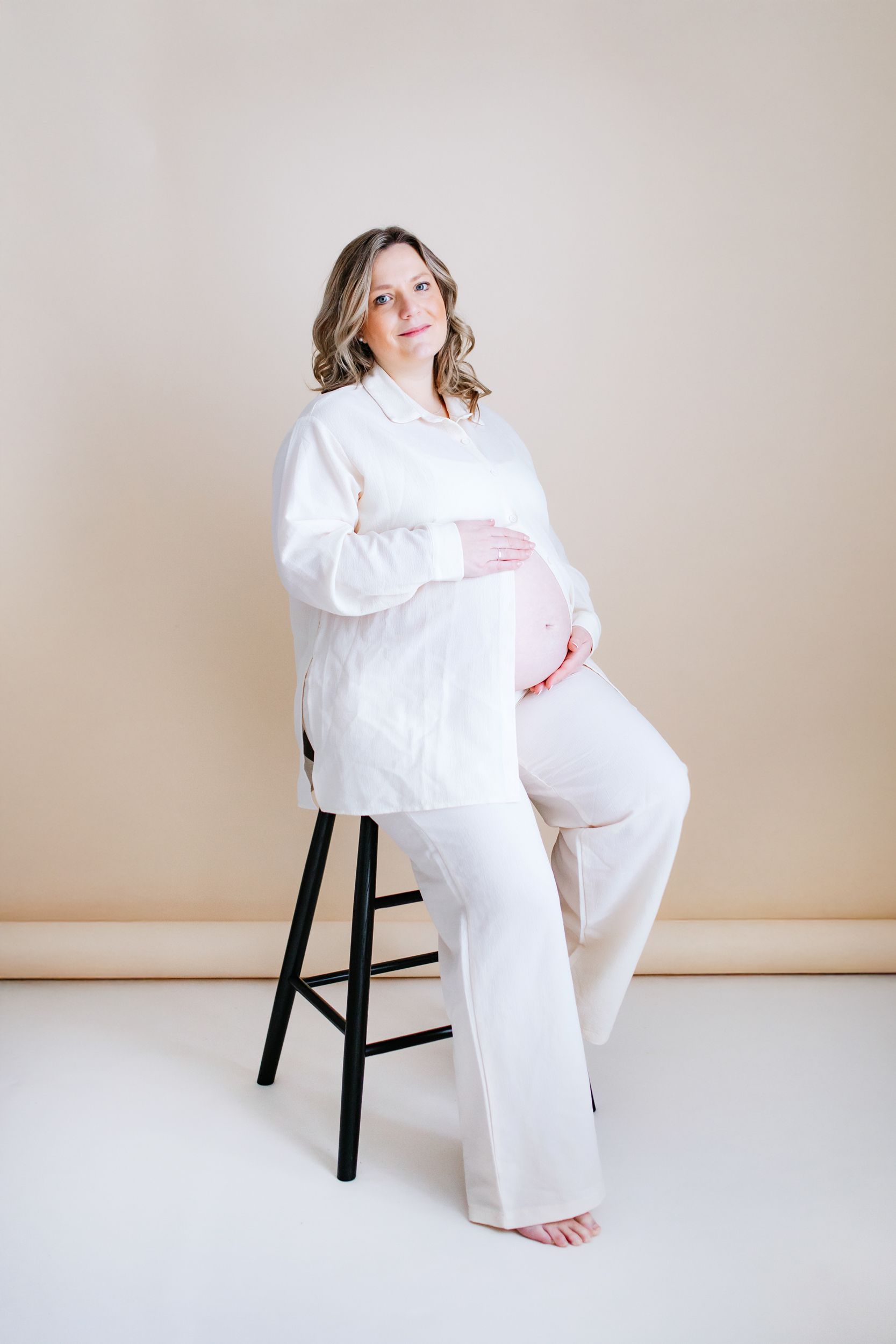 Pregnant woman in white clothing sits on a black stool against neutral backdrop during maternity photoshoot.