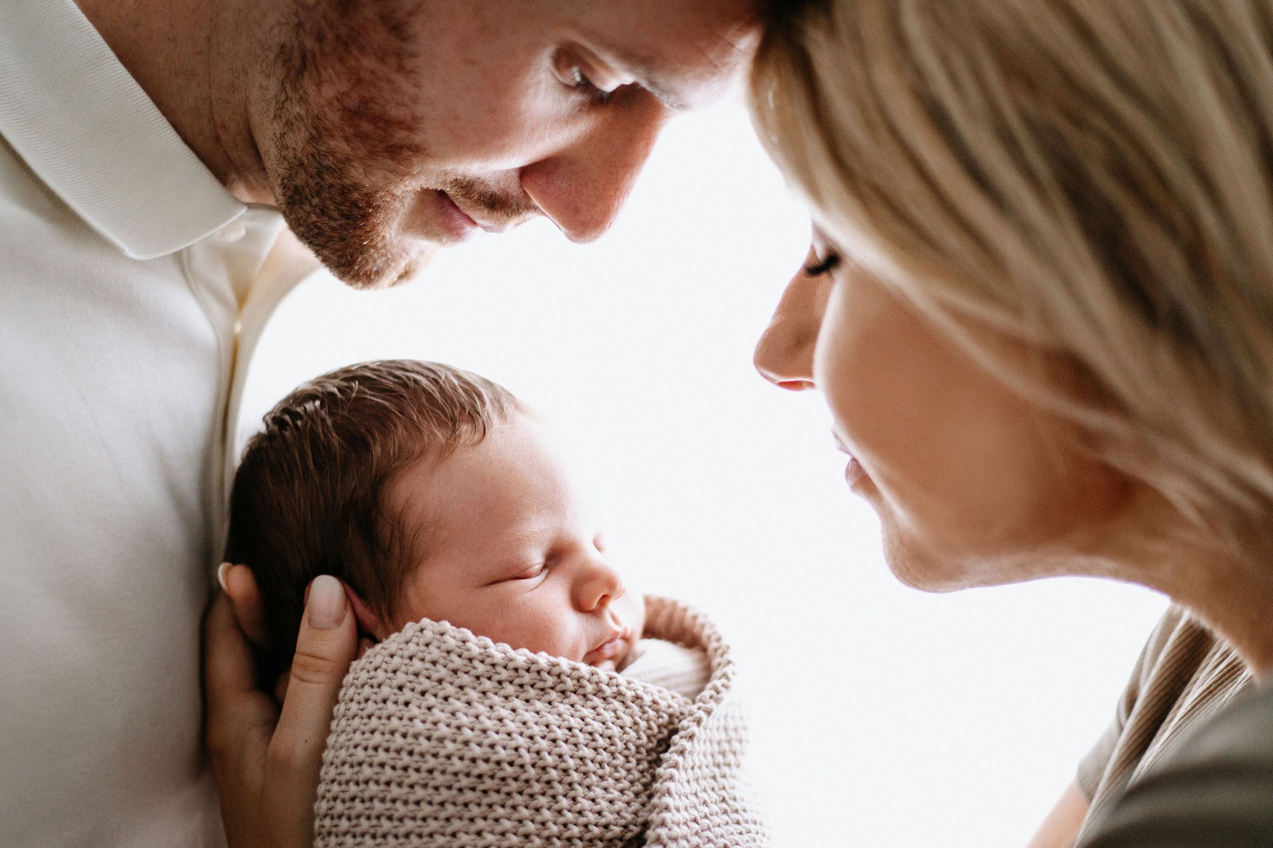 A tender family moment shows parents looking lovingly at their newborn baby wrapped in a knitted blanket.