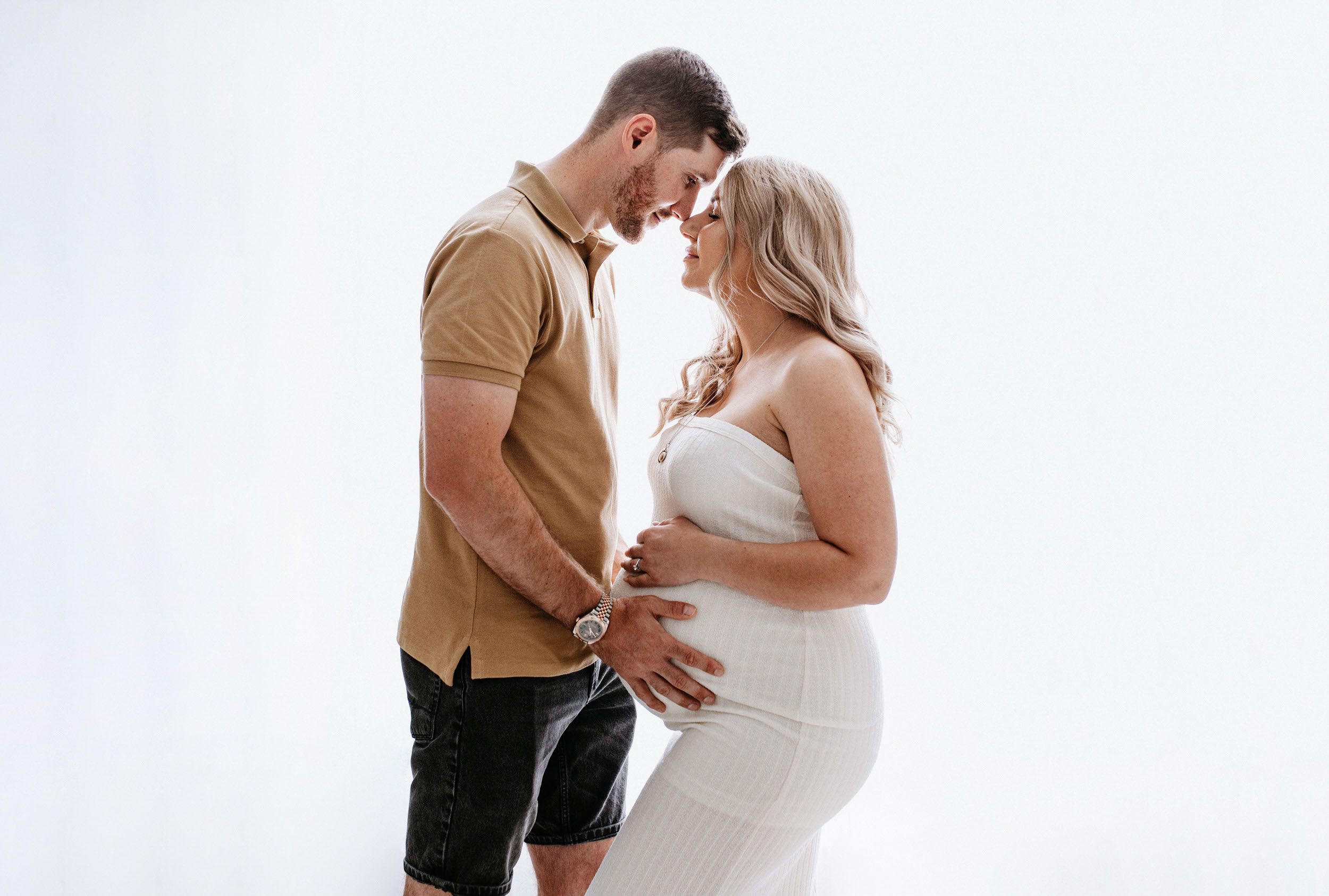 Intimate maternity photo with couple in white and tan standing close together against bright background.