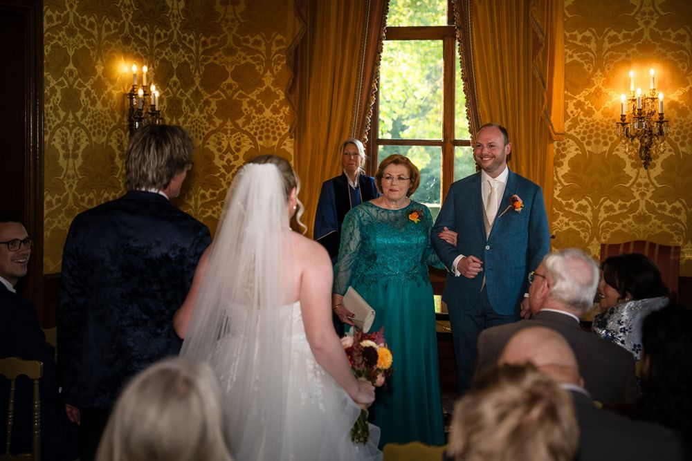 Een bruid wordt weggegeven door haar vader tijdens de trouwceremonie in kasteel Ruurlo. De bruidegom wacht haar lachend op met zijn moeder.