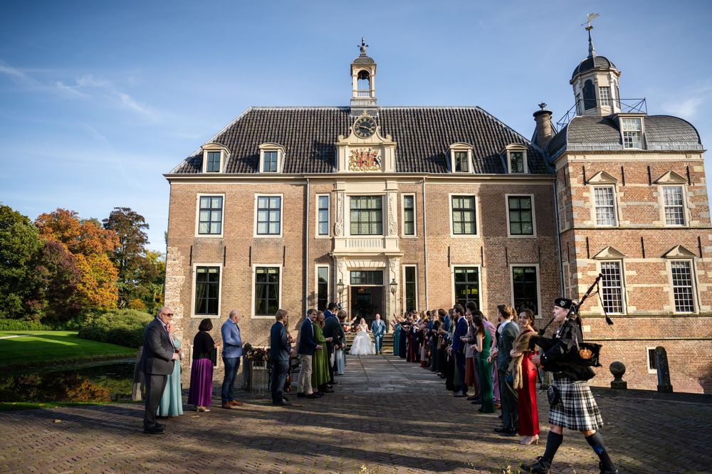 Bruidspaar loopt naar buiten na hun trouwceremonie in kasteel Ruurlo en wordt opgewacht door alle bruidsgasten en een doedelzakspeler.