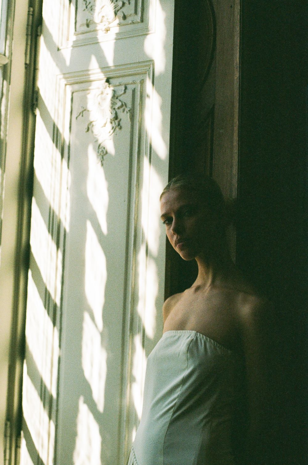 Sunlight streams through ornate window onto figure in white dress standing in shadowy corner of elegant room.