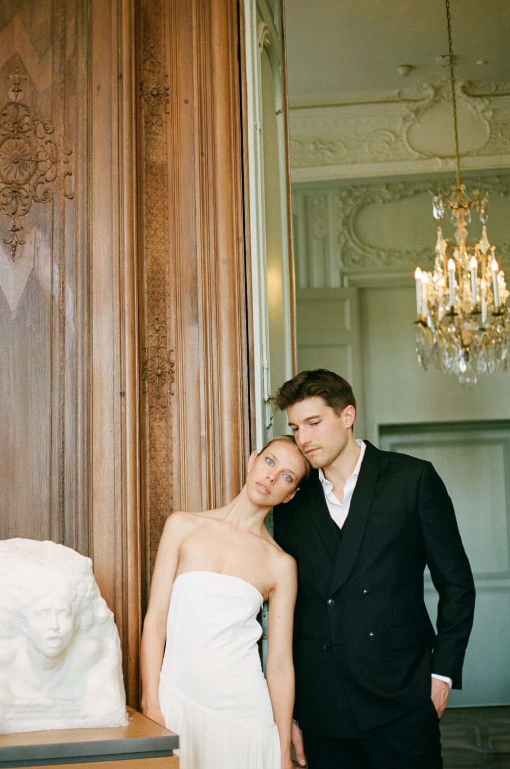 Couple in formal attire embraces in an elegant room with ornate wood paneling and crystal chandelier.