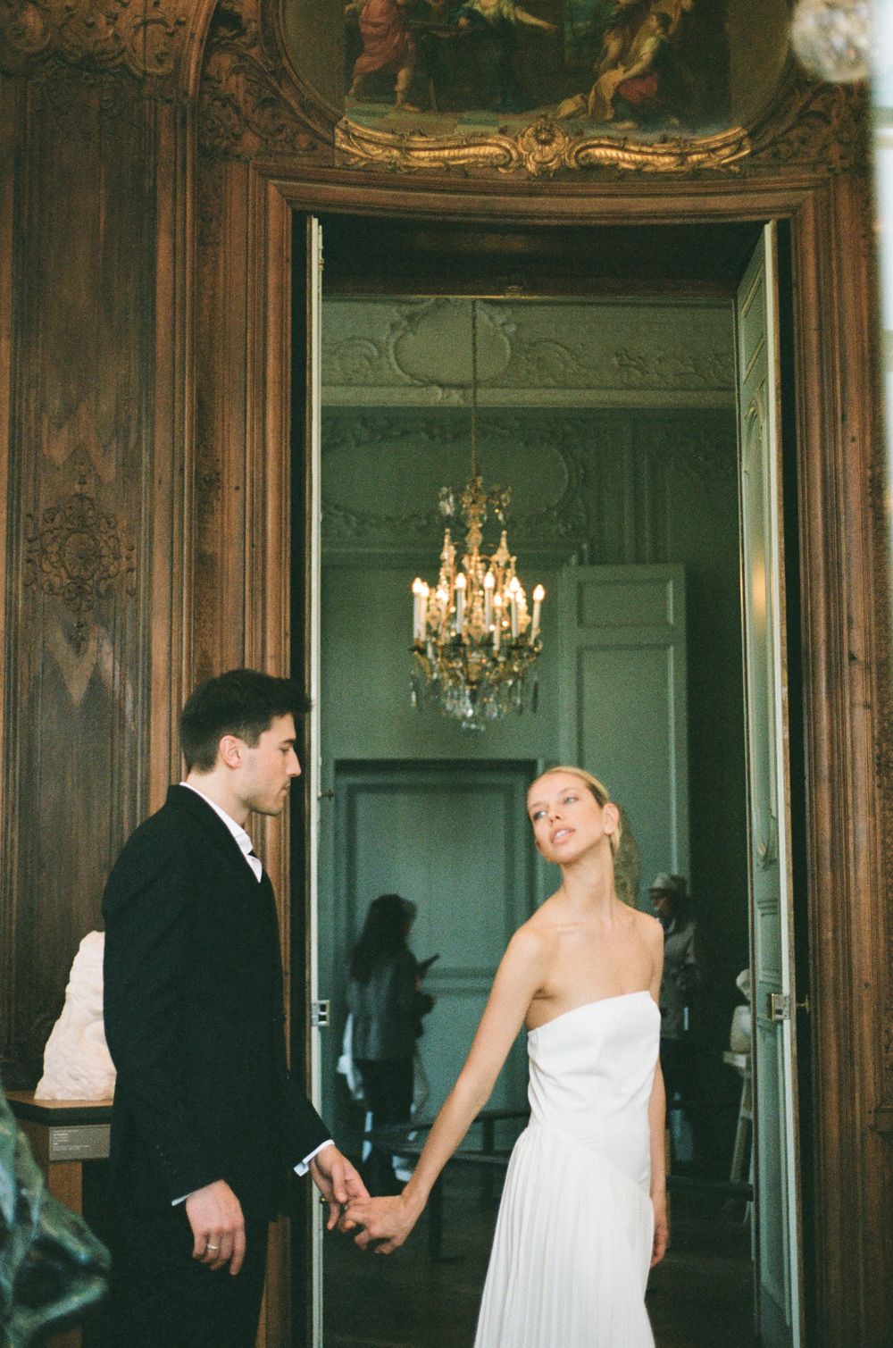 A couple holds hands in an elegant room with ornate wooden doorframe and crystal chandelier in the background.