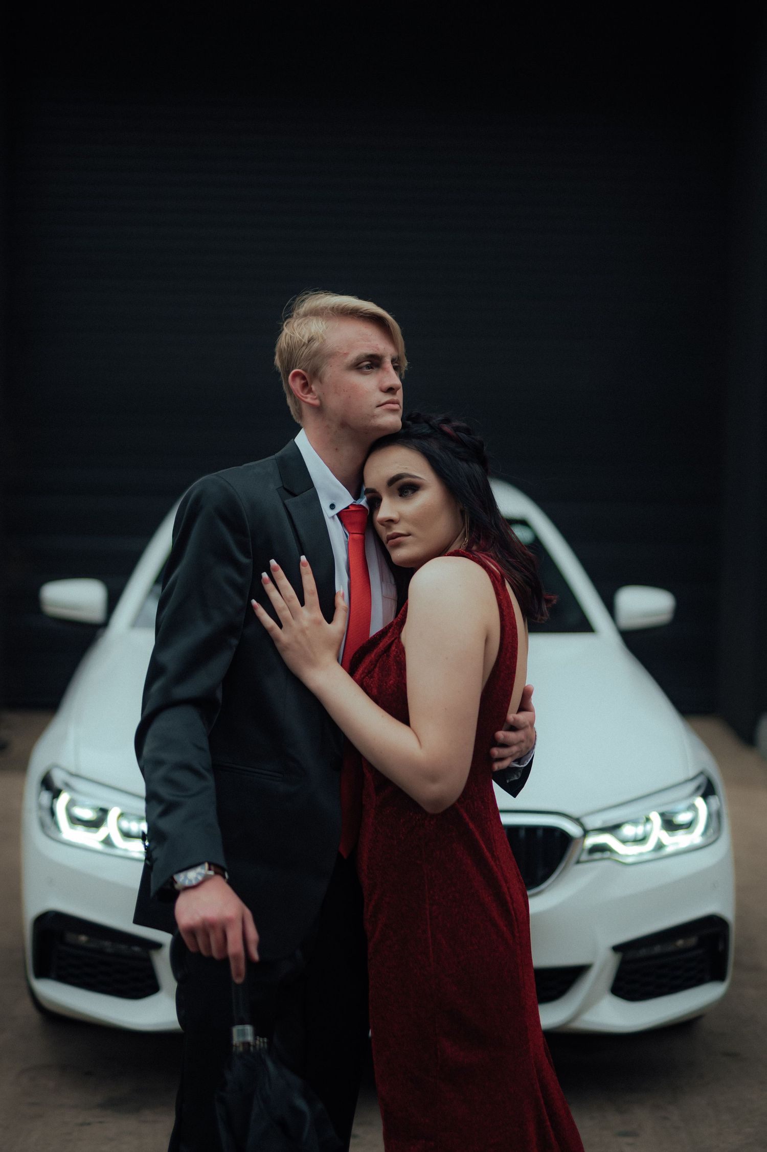 A glamorous couple in formal attire posing in front of a white luxury car.