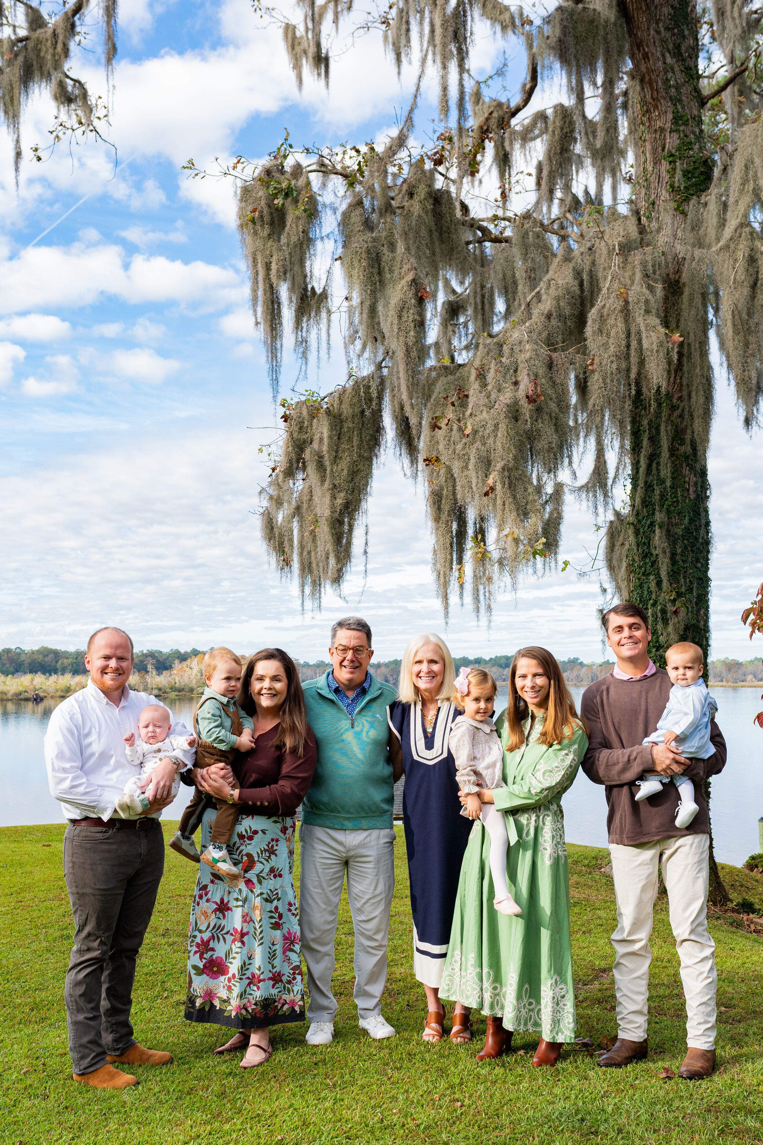 Extended family group poses together outdoors under Spanish moss trees near a lake on a sunny day.