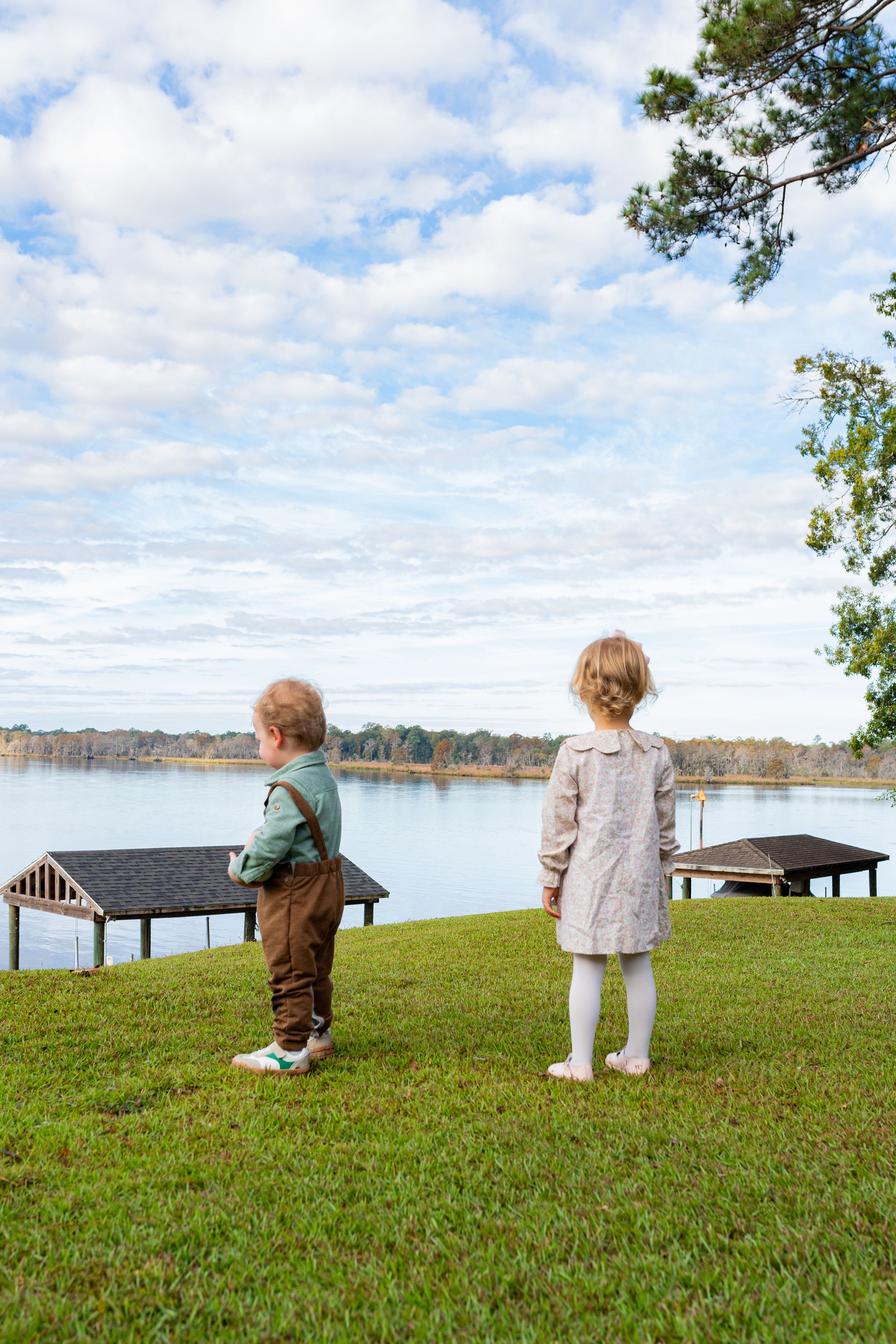 Two children stand on grassy hill overlooking a lake with boat docks and cloudy skies in the background.