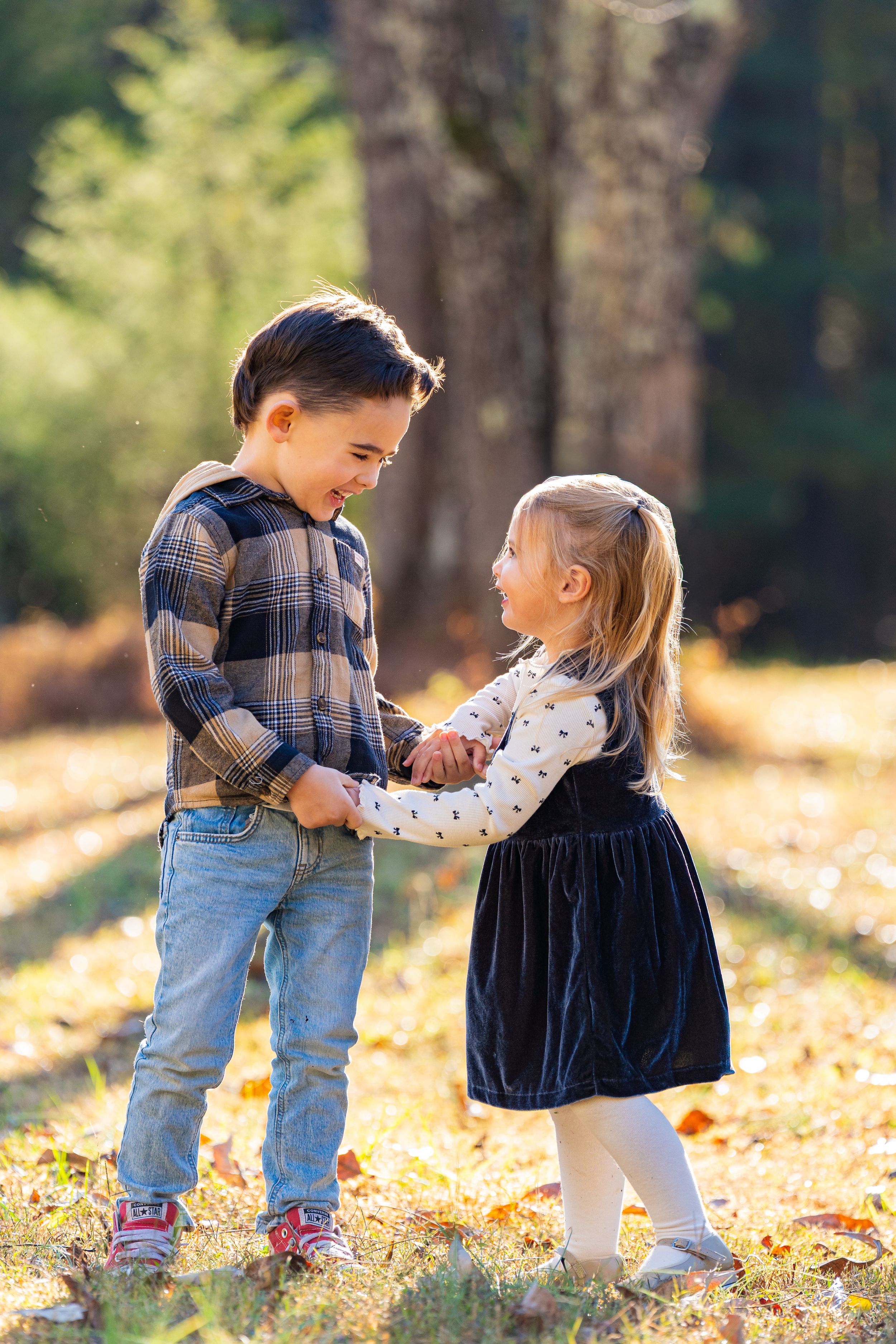 Two toddlers play together in autumn leaves wearing plaid and denim outfits in a sunlit forest setting.
