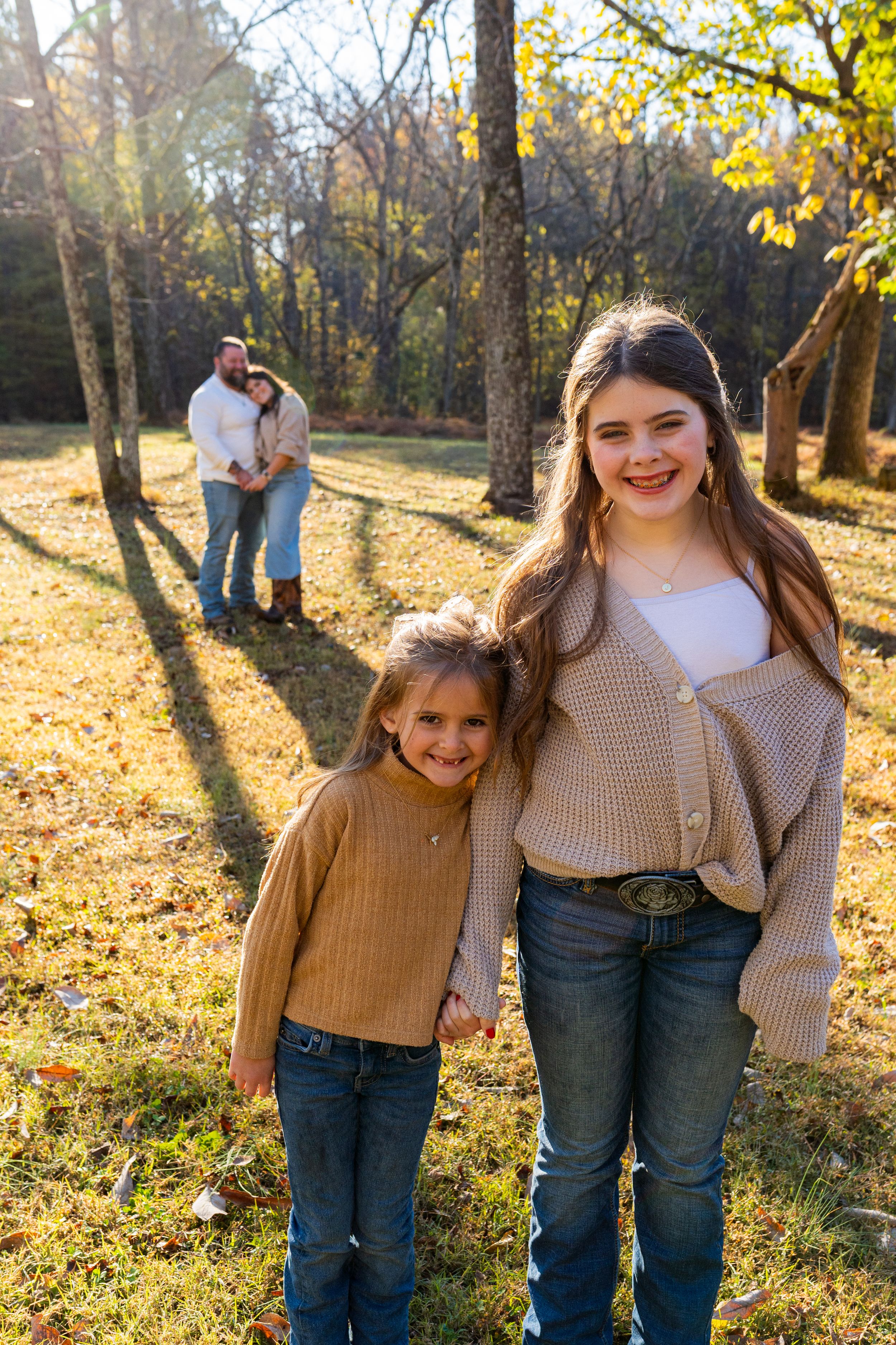 Two people in casual fall attire share joyful moments in a sunlit autumn forest with golden leaves scattered on the ground.