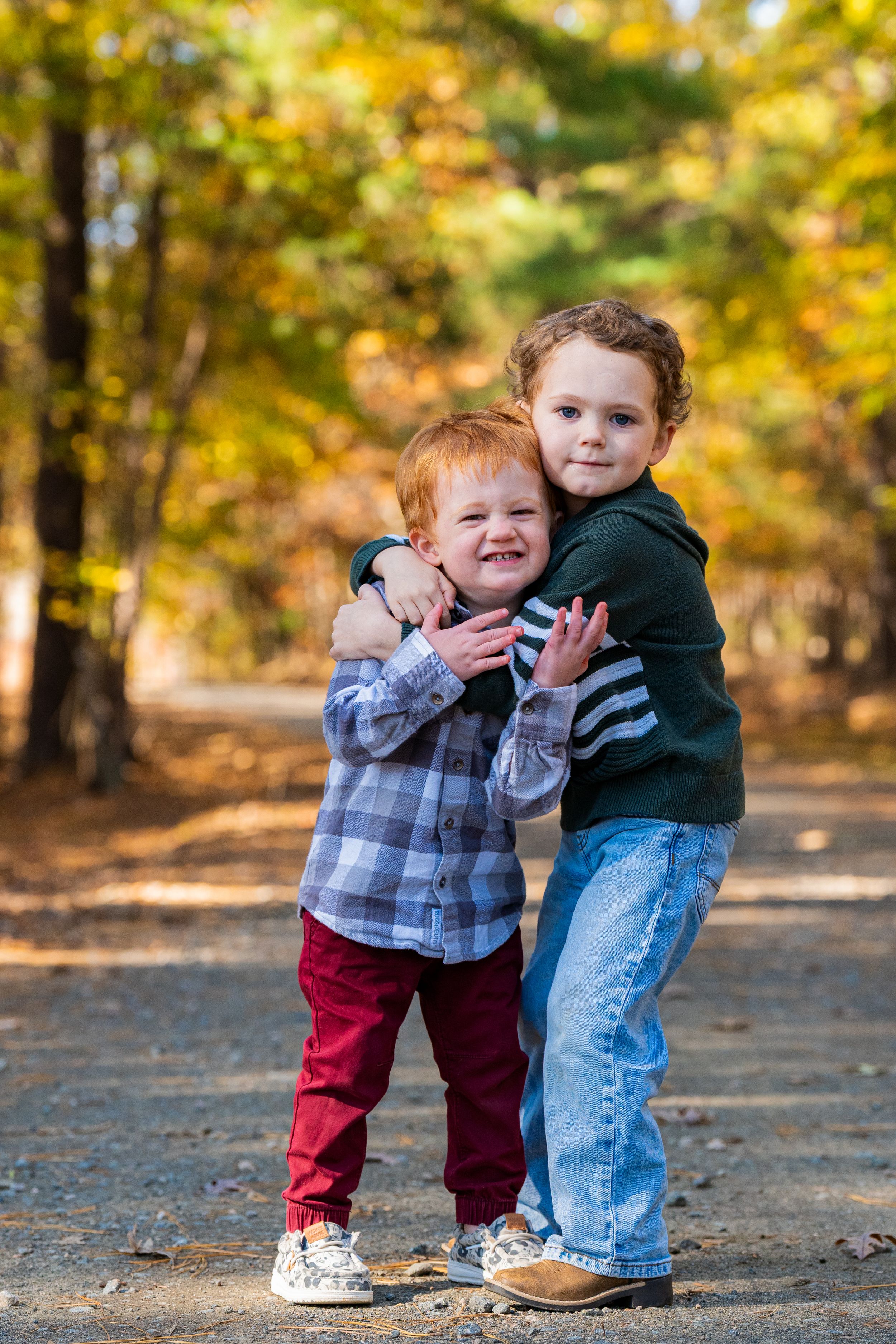 Two young children share a sweet hug outdoors surrounded by autumn foliage and fallen leaves.