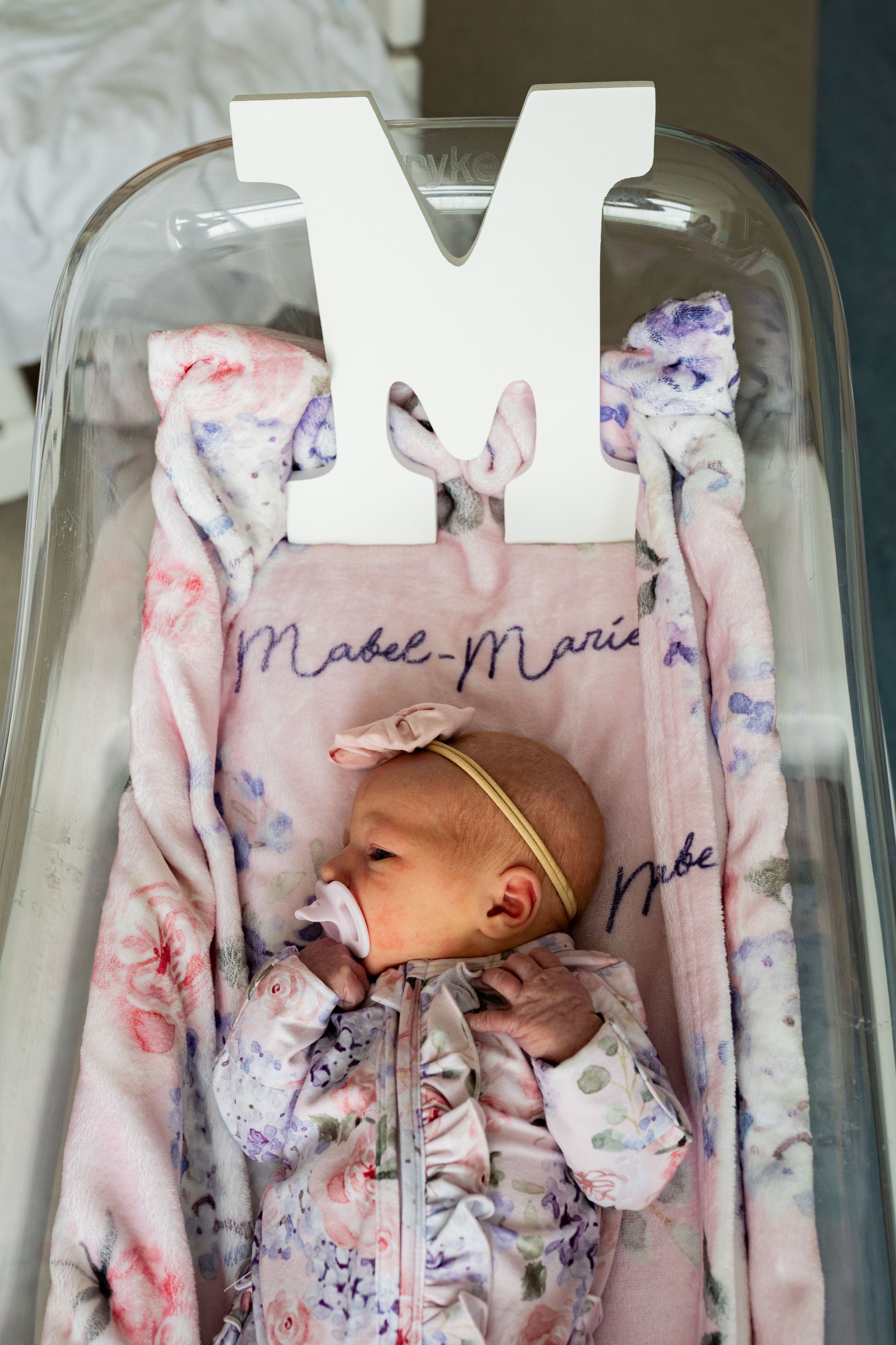 Clear plastic baby bassinet with a white decorative 'M' letter and floral pink and purple patterned blanket inside.
