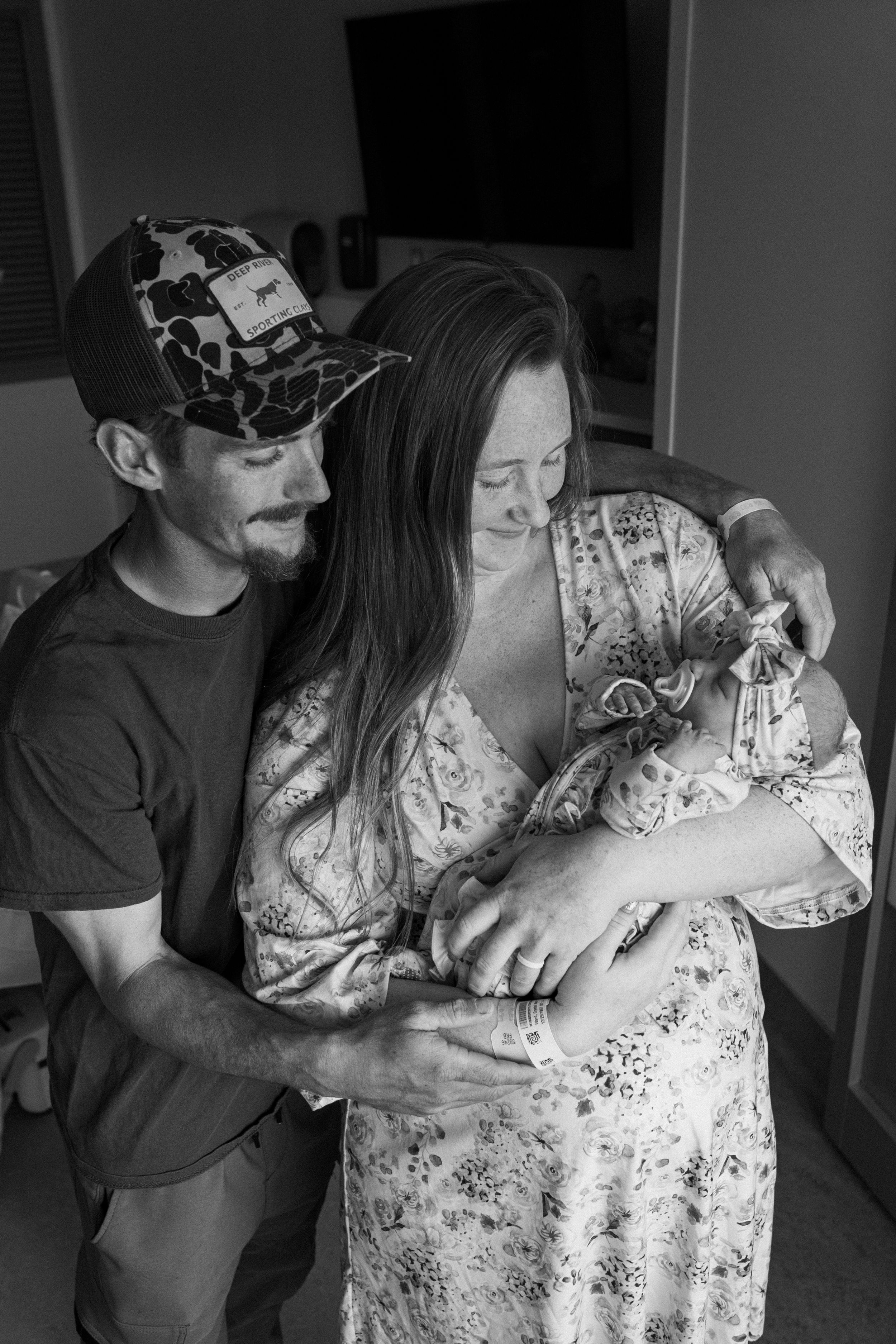 A tender black and white moment captured as couple embrace while holding their newborn baby in a floral dress.