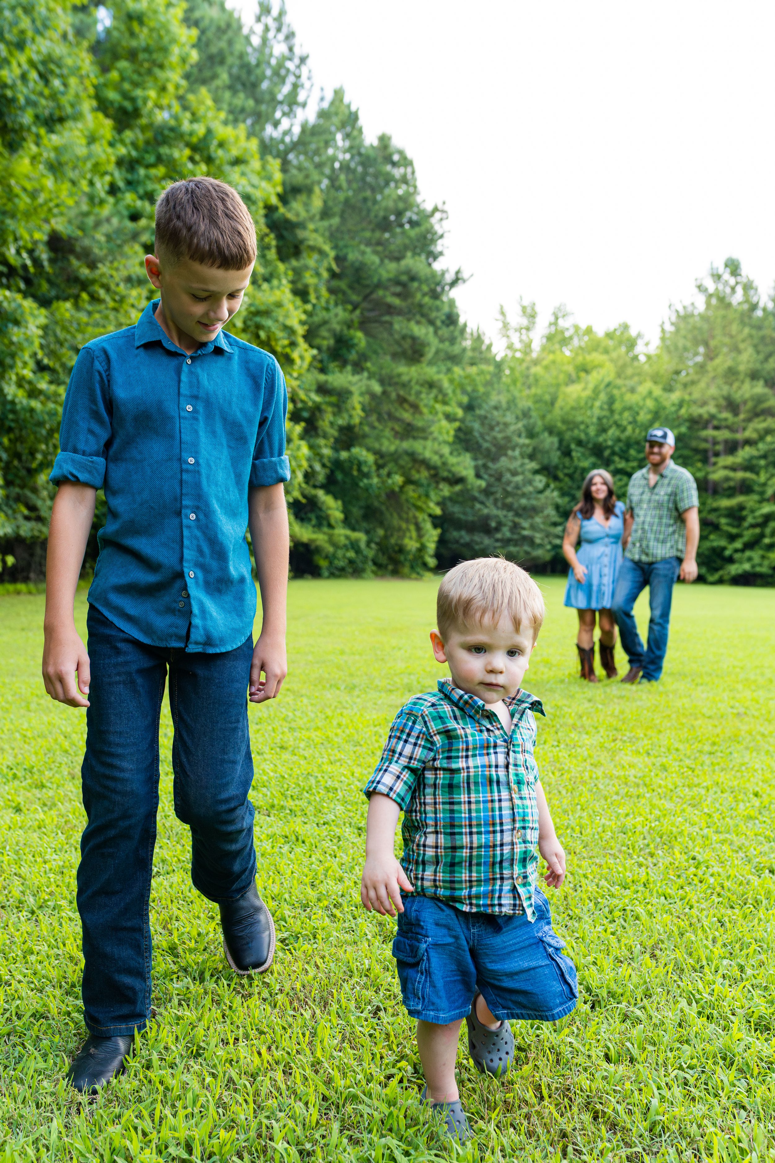 Children playing in a grassy field on a sunny day with adults standing in the background near trees.