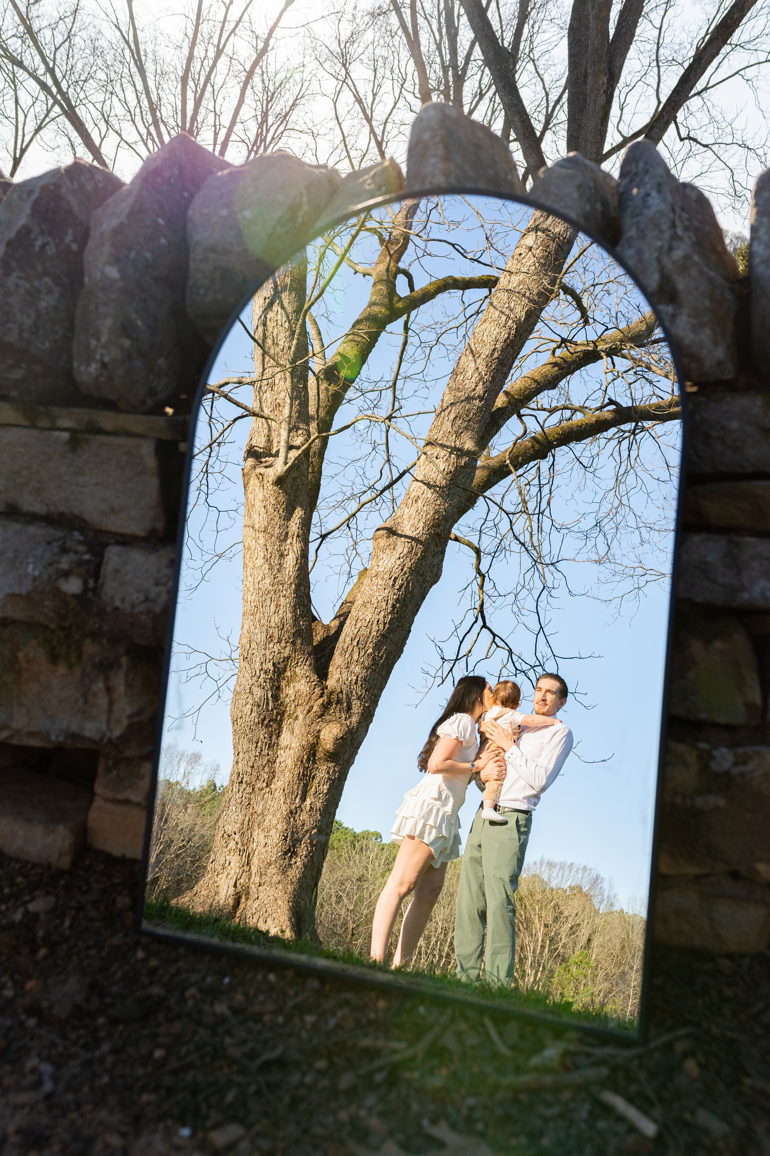 Stone archway frames a romantic outdoor scene with a couple standing beneath a large bare tree against blue sky.