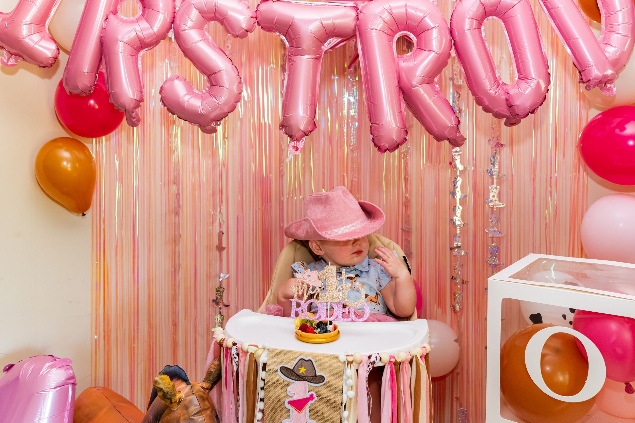 A young girl in a pink cowboy hat sits at a decorated table with pink balloons and streamers at a birthday party.