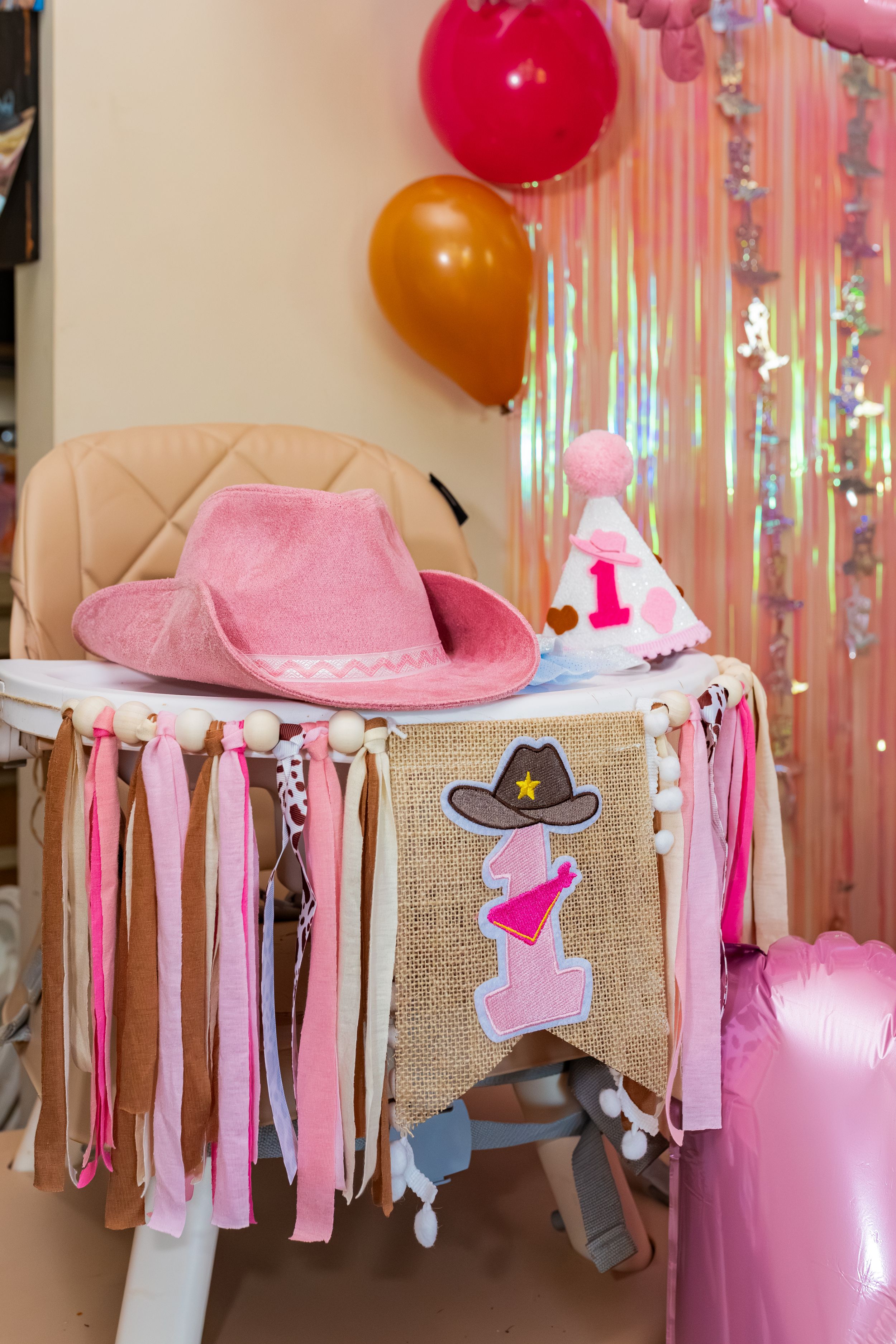 Pink cowboy hat on a decorated first birthday high chair with balloons and colorful streamers in the background.