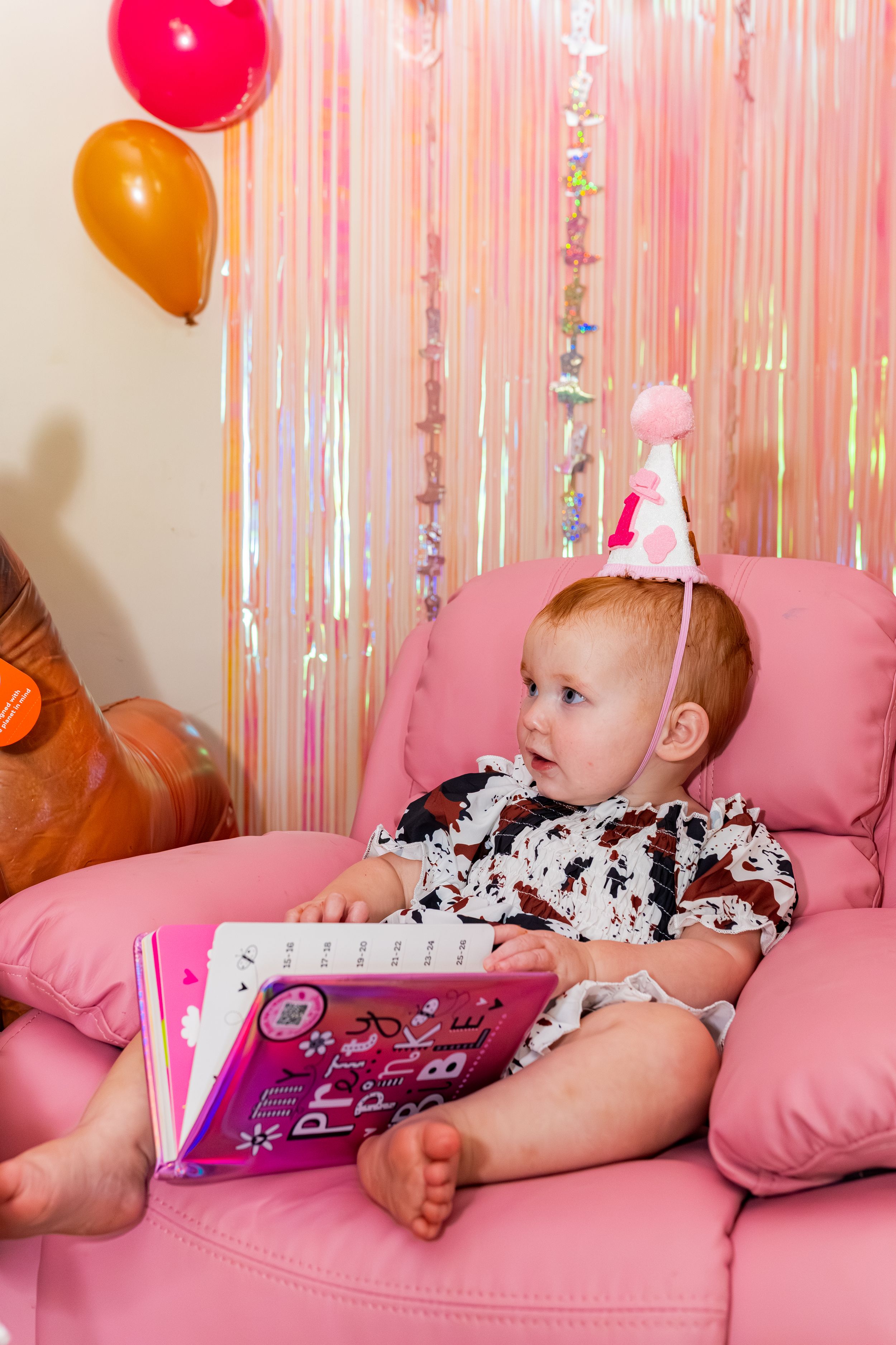 Baby girl in pink party hat sits in pink chair holding a book, surrounded by colorful balloons and shimmery streamers.