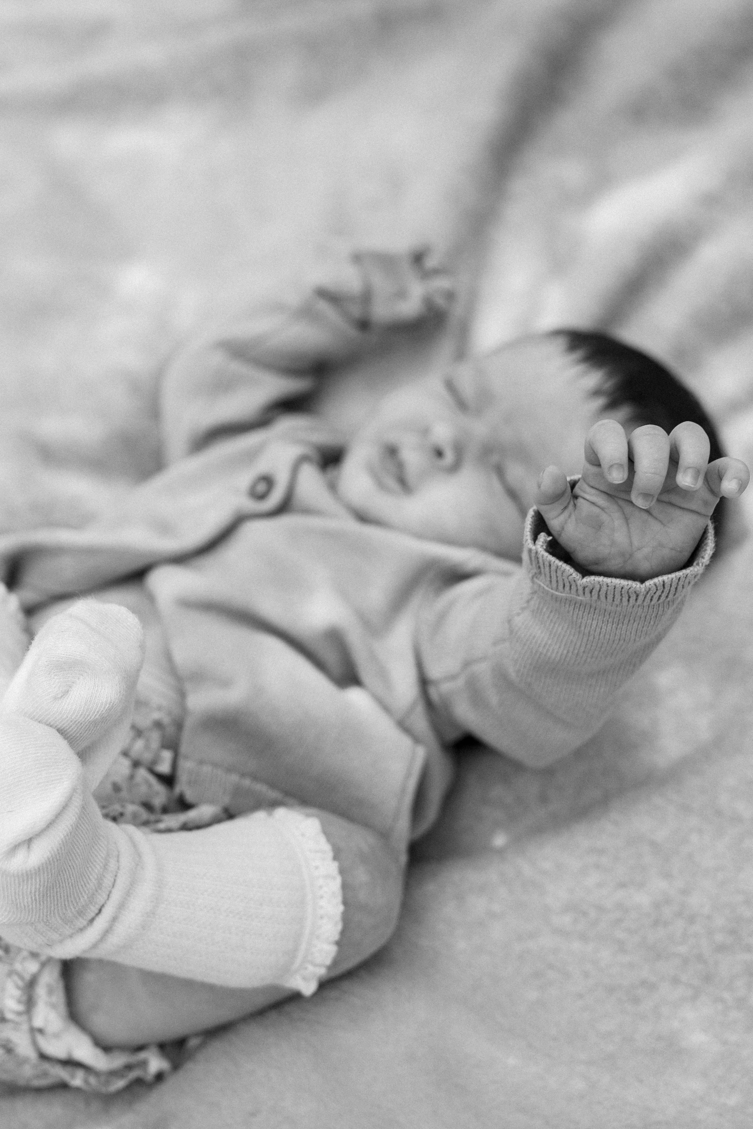 Black and white photo of a newborn baby lying down, wearing a onesie and socks, with tiny hand raised.