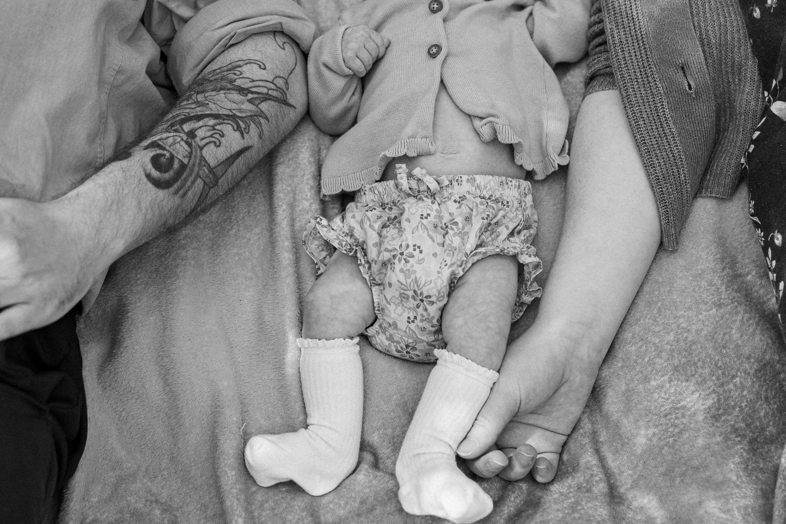 Black and white photo of a newborn baby in floral bloomers and white socks held by parents, one with a tattoo.